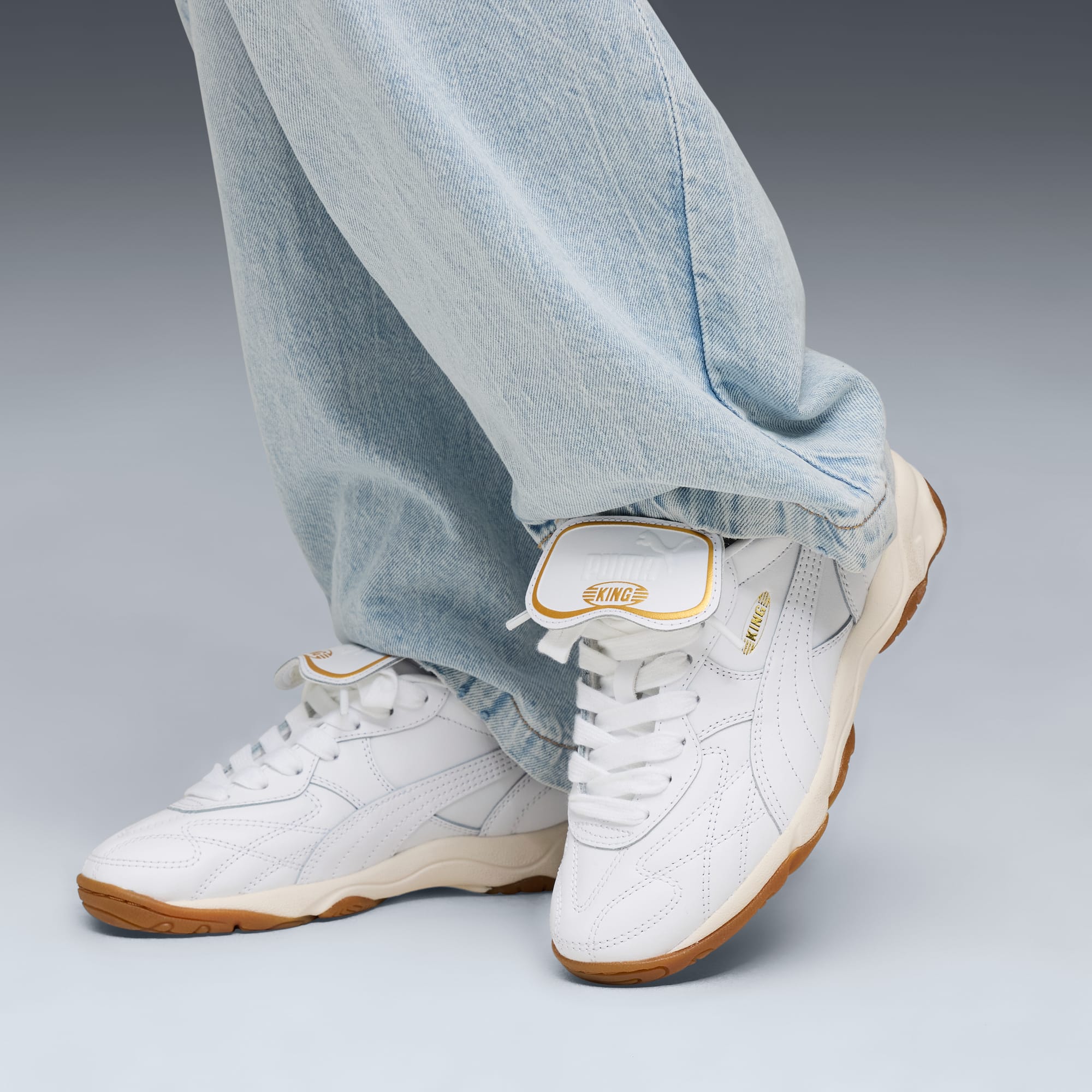 Against grey, a person wears light jeans and white leather trainers with tan soles.
