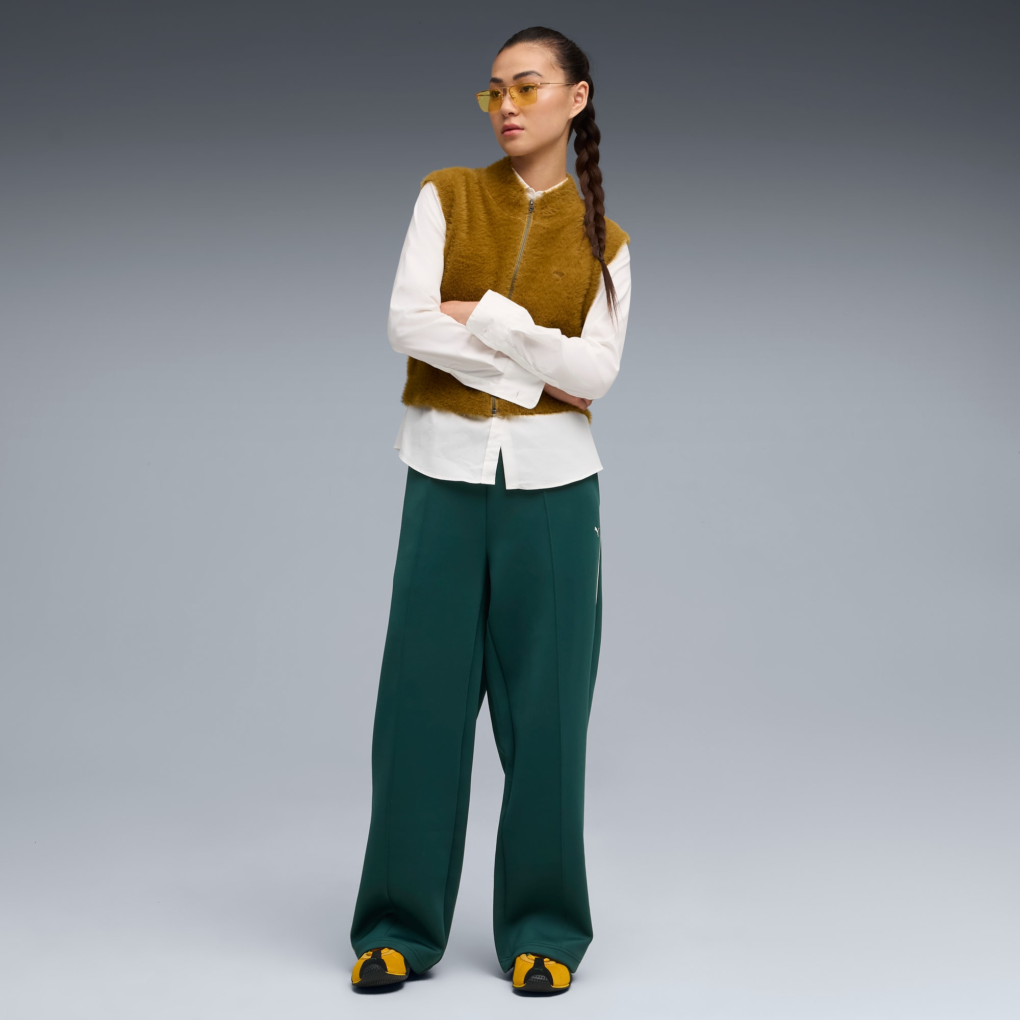 Sur fond gris, un mannequin aux bras croisés porte un gilet marron, une chemise blanche, un pantalon vert et des lunettes jaunes.