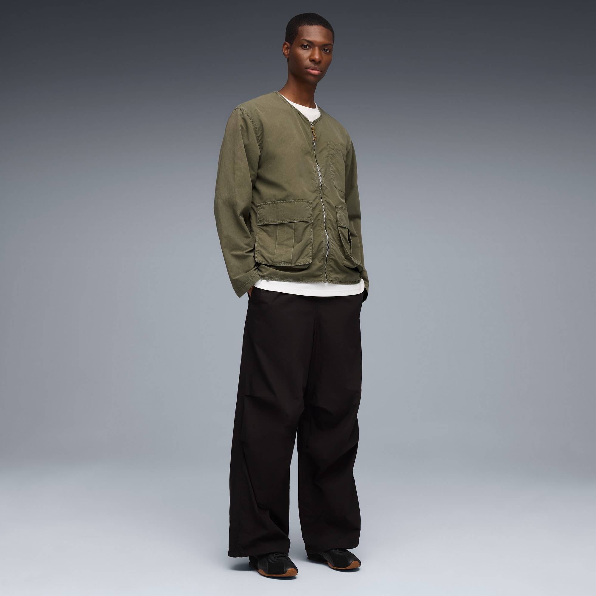 Un mannequin pose sur fond gris, vêtu d'une veste vert clair, d'un t-shirt blanc et d'un pantalon noir.