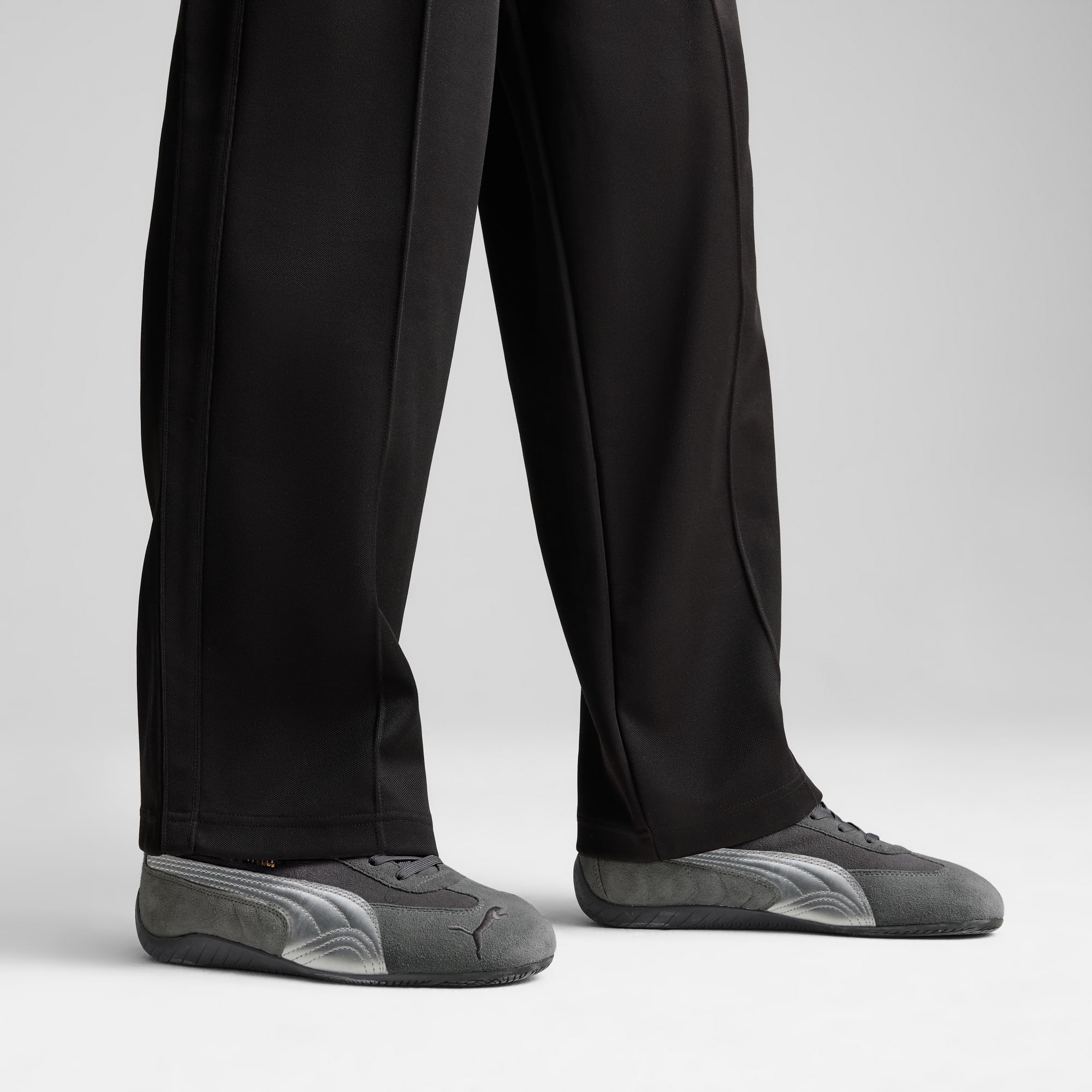Sur fond gris, des jambes en pantalon noir et baskets grises à détails argentés sont visibles.
