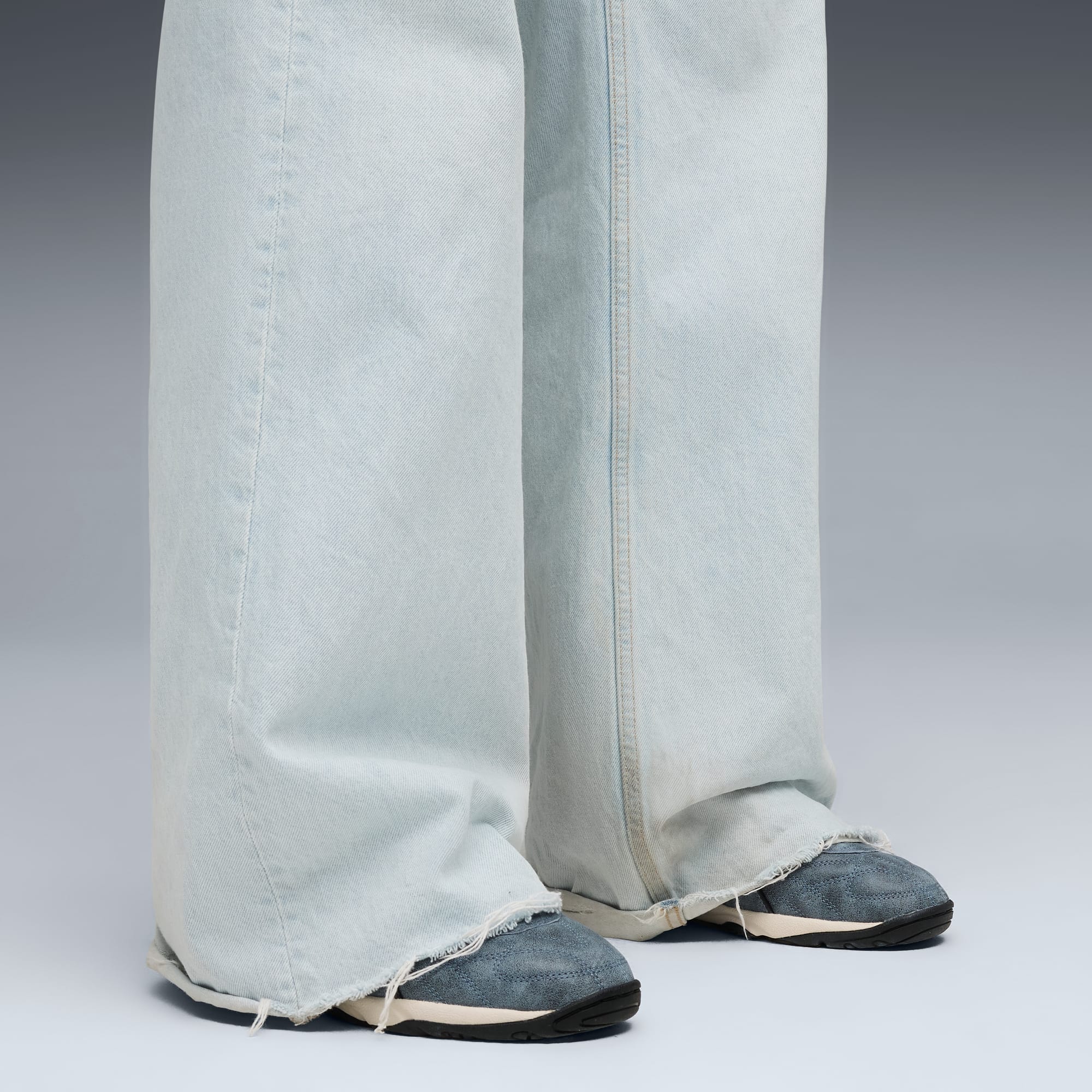 Tegen een grijze achtergrond draagt een model een lichtblauwe jeans met gerafelde zoom en blauwe sneakers.