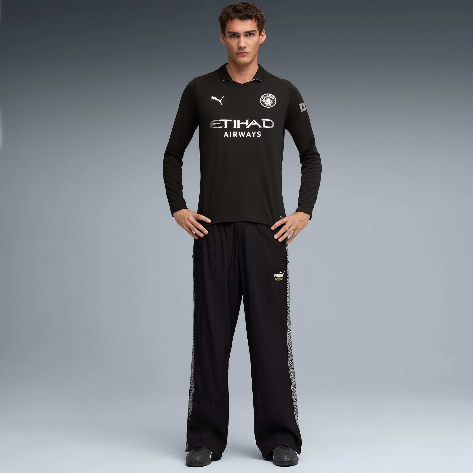 Tegen een grijze achtergrond poseert een model in een zwart shirt met lange mouwen en broek.