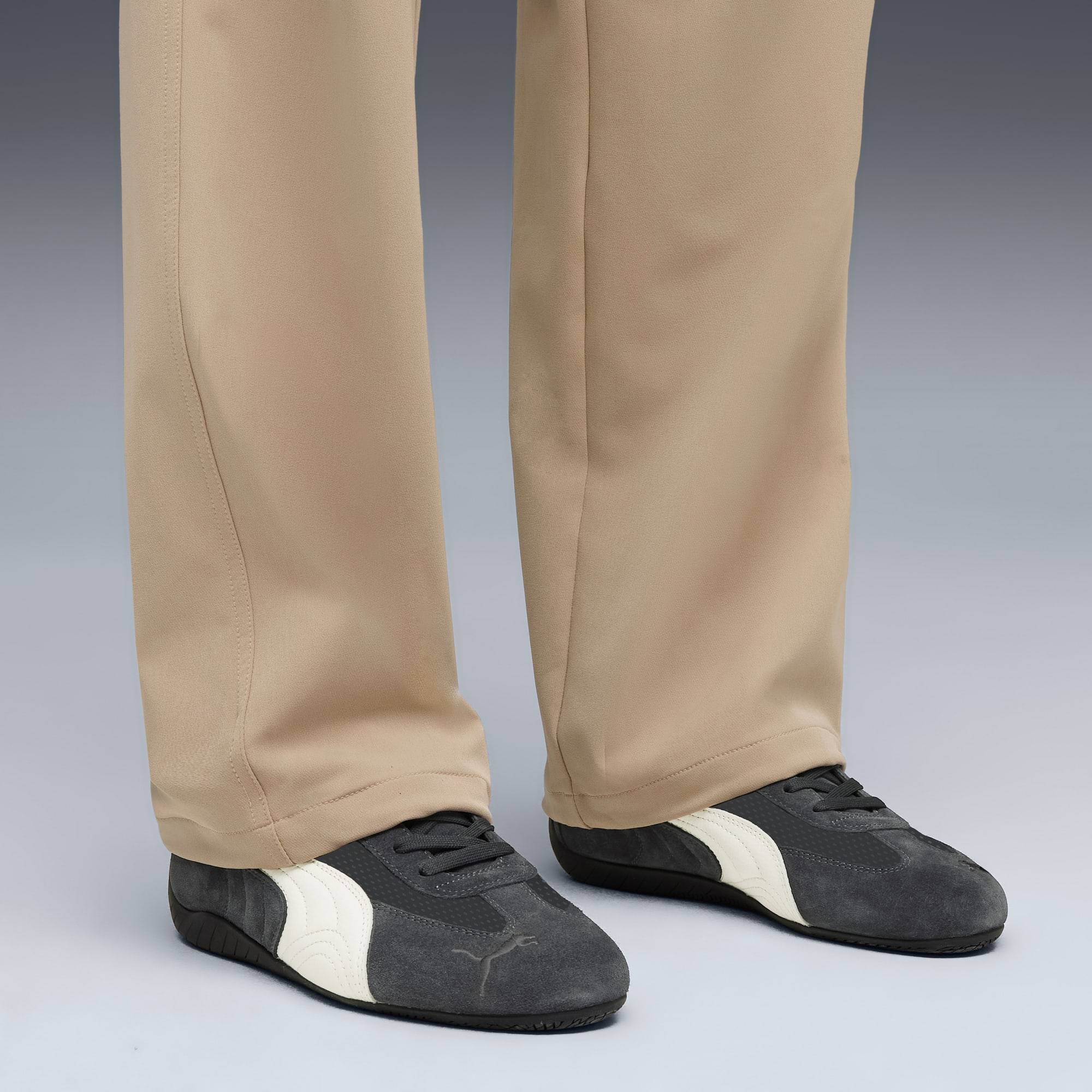 Tegen een grijze achtergrond draagt een model een beige broek en zwarte sneakers met witte strepen.