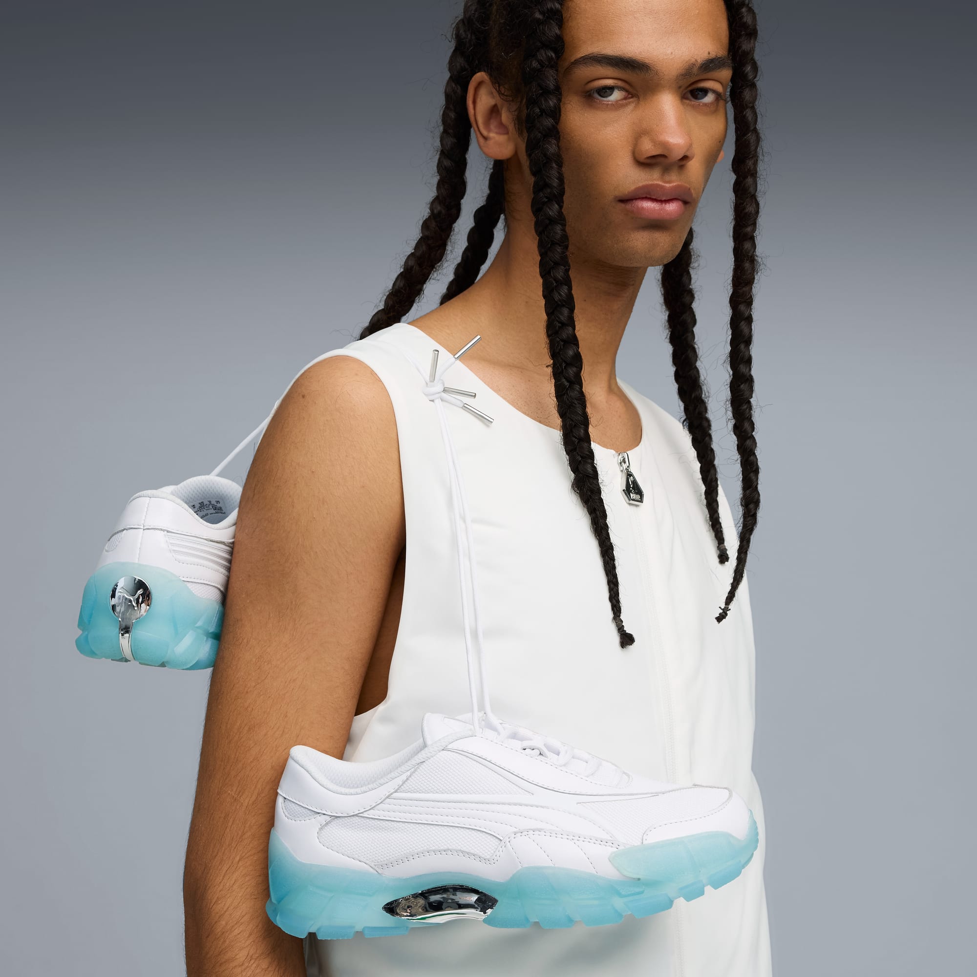 Sur fond gris, un mannequin avec des tresses porte un haut blanc et des baskets blanches.