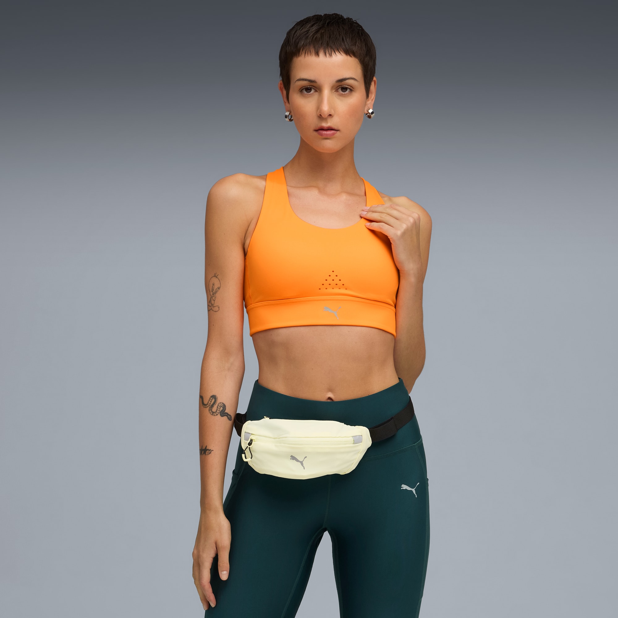 Sur fond gris, un mannequin pose avec un haut orange, legging turquoise et banane claire.