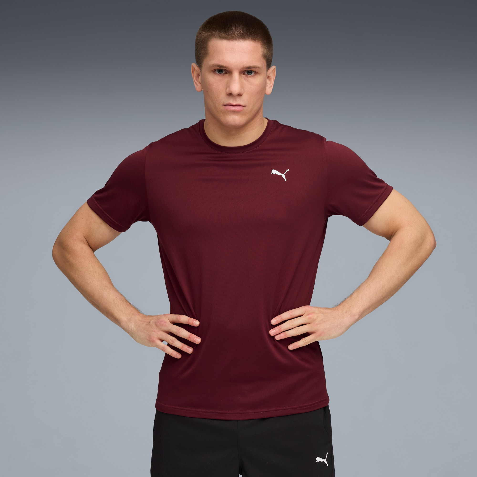 Tegen een grijze achtergrond poseert een model met de handen op de heupen, in bordeauxrood shirt en zwarte short.