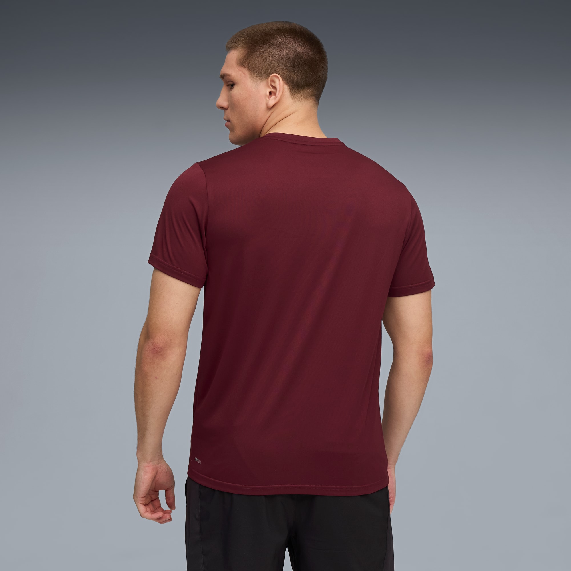 Een model staat met de rug naar de camera op een grijze achtergrond, met een bordeauxrood shirt en zwarte korte broek.