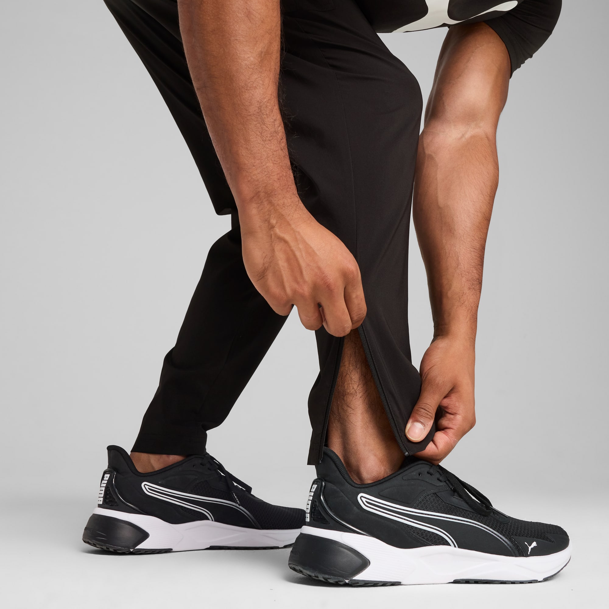 Sur fond gris, une personne ajuste la fermeture éclair d'un pantalon noir avec des baskets noires et blanches.