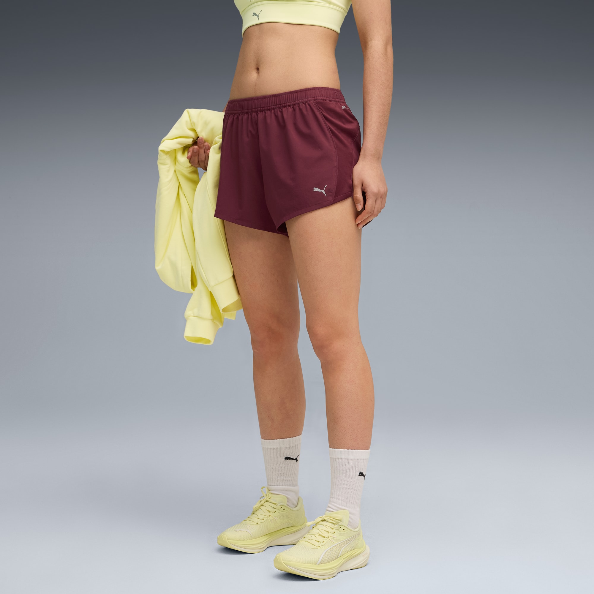 Un modèle pose sur fond gris, portant un haut jaune, un short bordeaux et des chaussures de course jaunes.