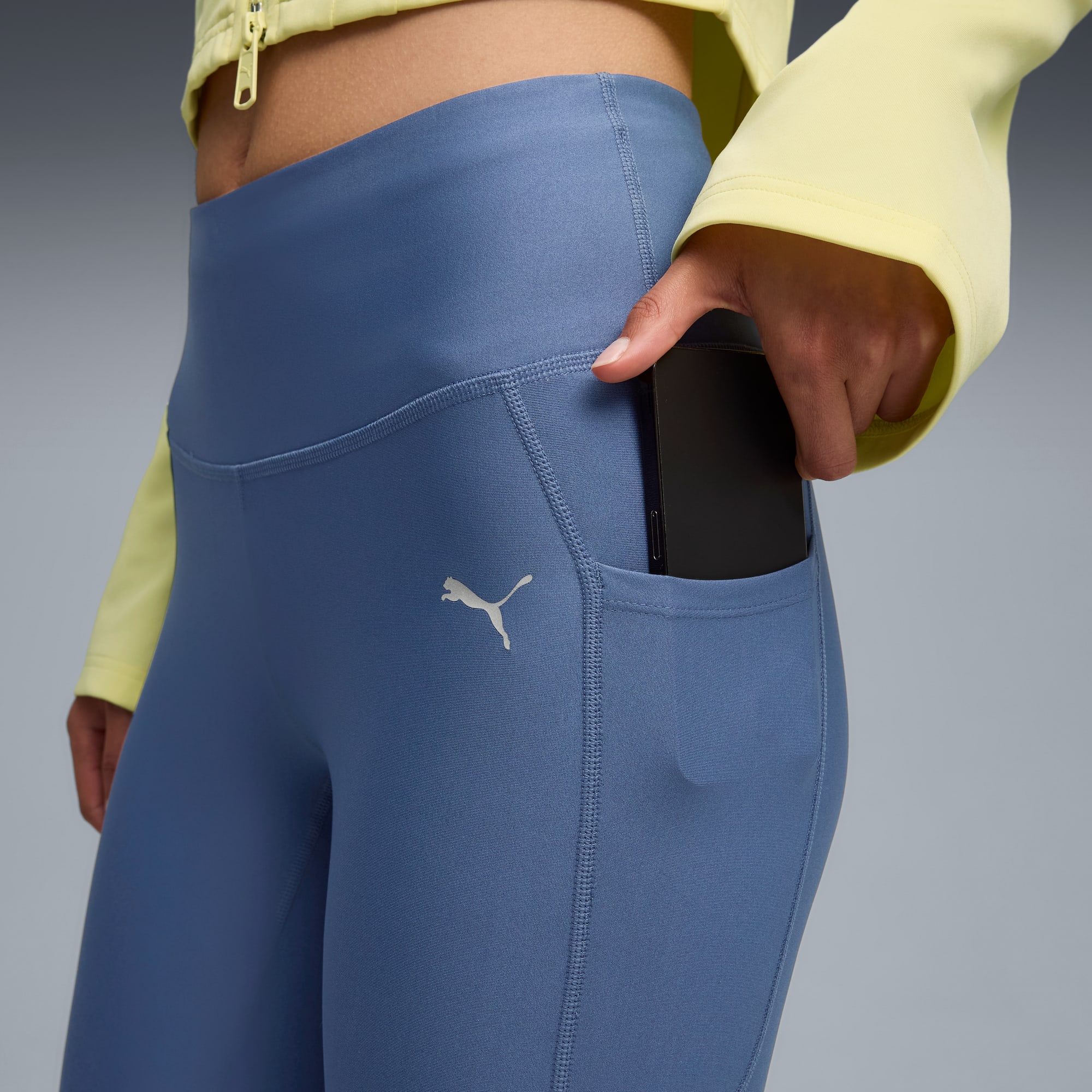 Sur fond gris, un mannequin glisse un téléphone dans la poche d'un legging bleu.