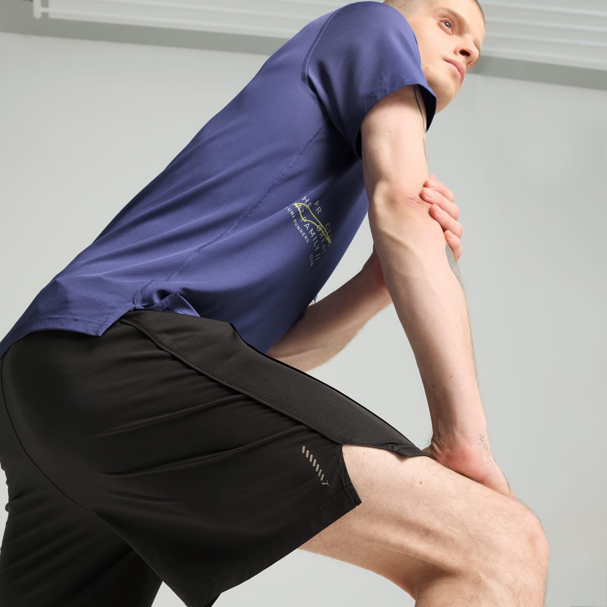 Een model rekt voor een grijze achtergrond, met een blauw shirt en zwarte shorts.