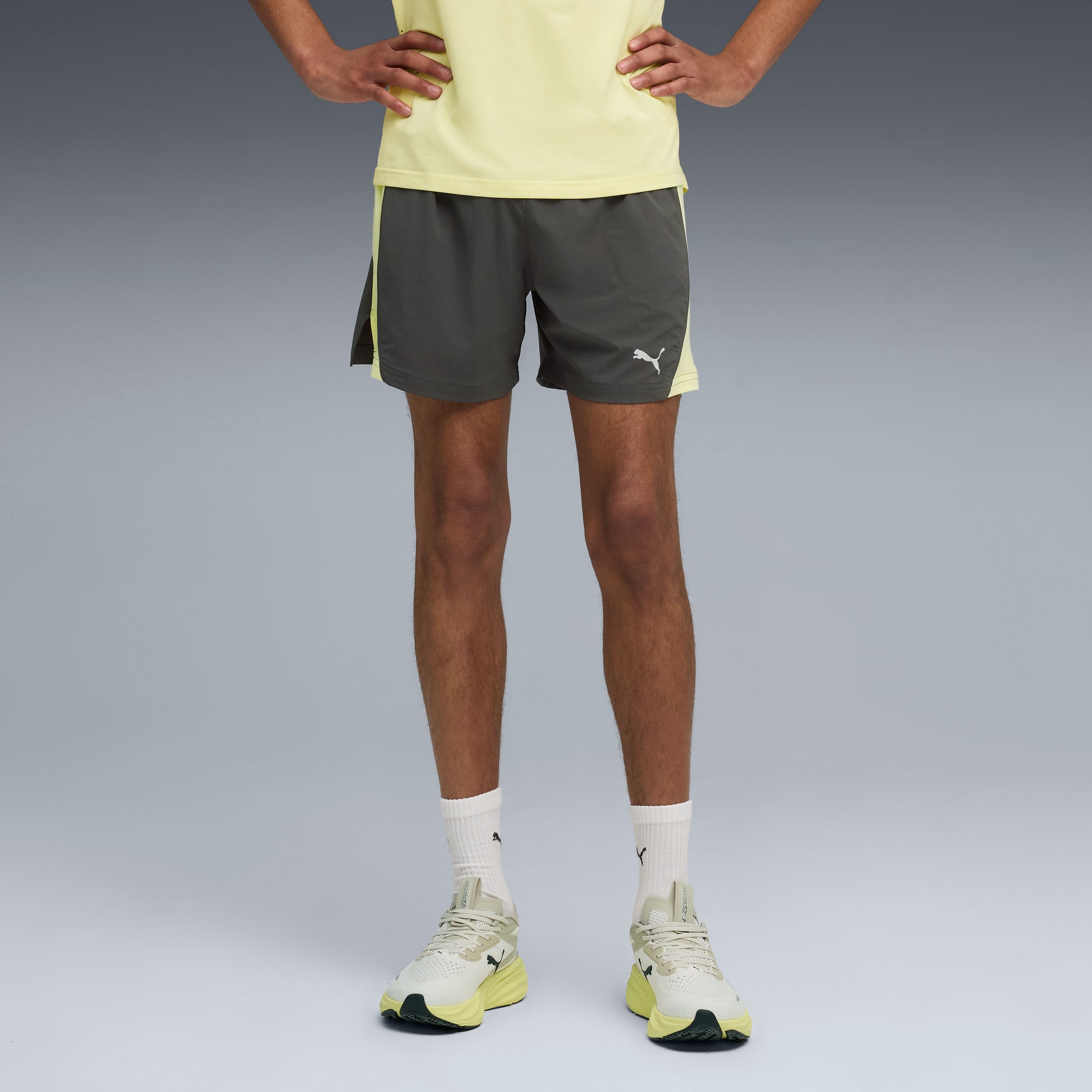 Op grijs, een model poseert in een gele top, grijze short met logo, sokken en hardloopschoenen.