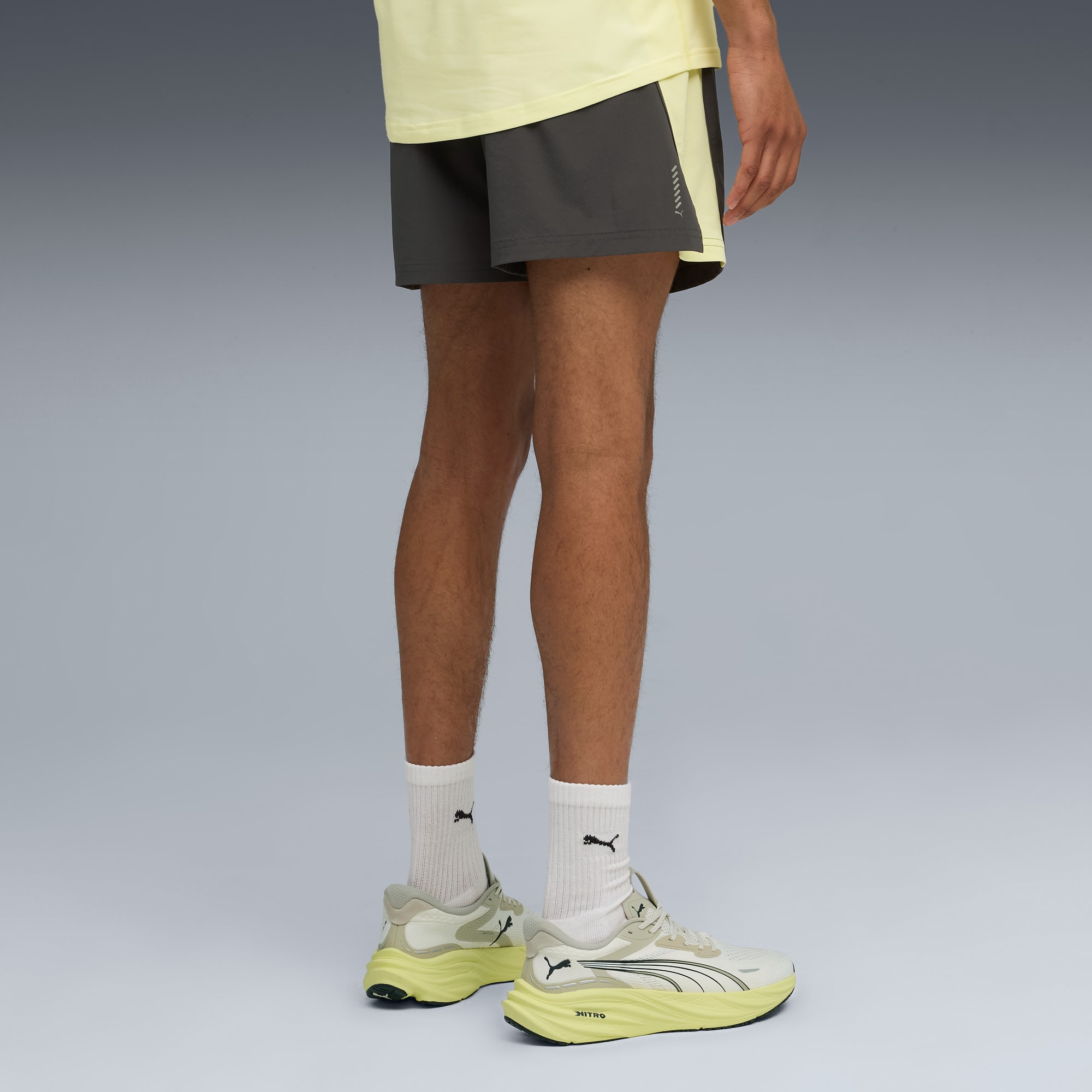 Een model poseert tegen grijs met een gele top, grijze korte broek en hardloopschoenen.