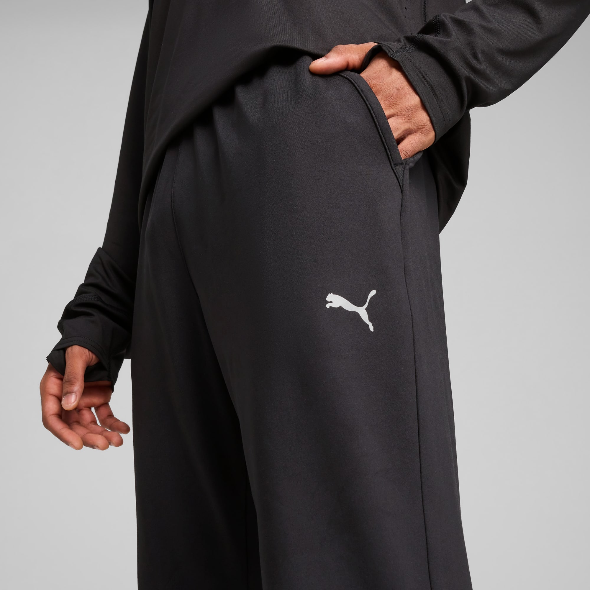 Vor grauem Hintergrund steckt ein Model die Hand in die Tasche einer schwarzen Laufhose mit Logo.
