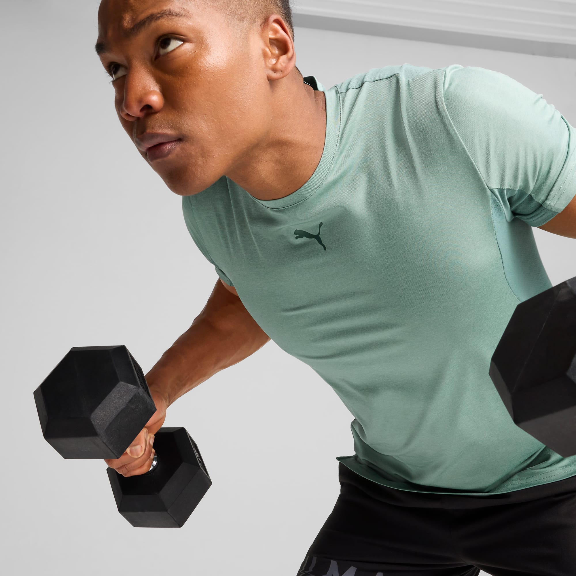 Tegen een grijze achtergrond heft een model gewichten, gekleed in een groen t-shirt en zwarte shorts.