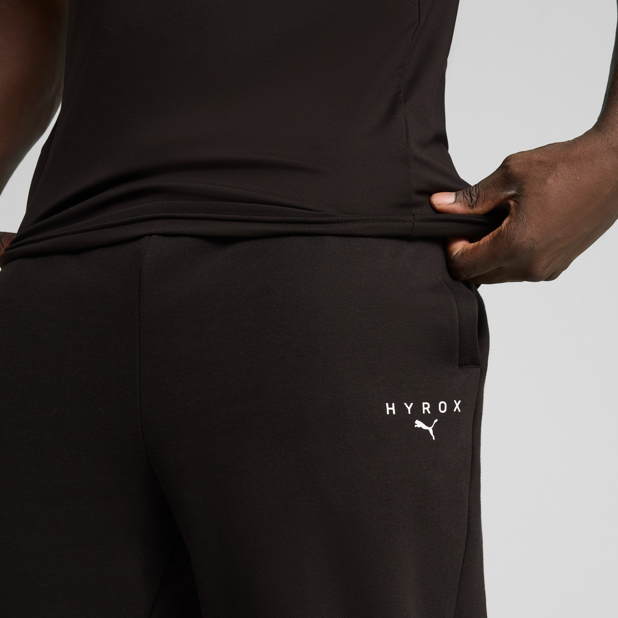 Op grijs houdt een model een zwarte HYROX broek en een zwarte top vast.