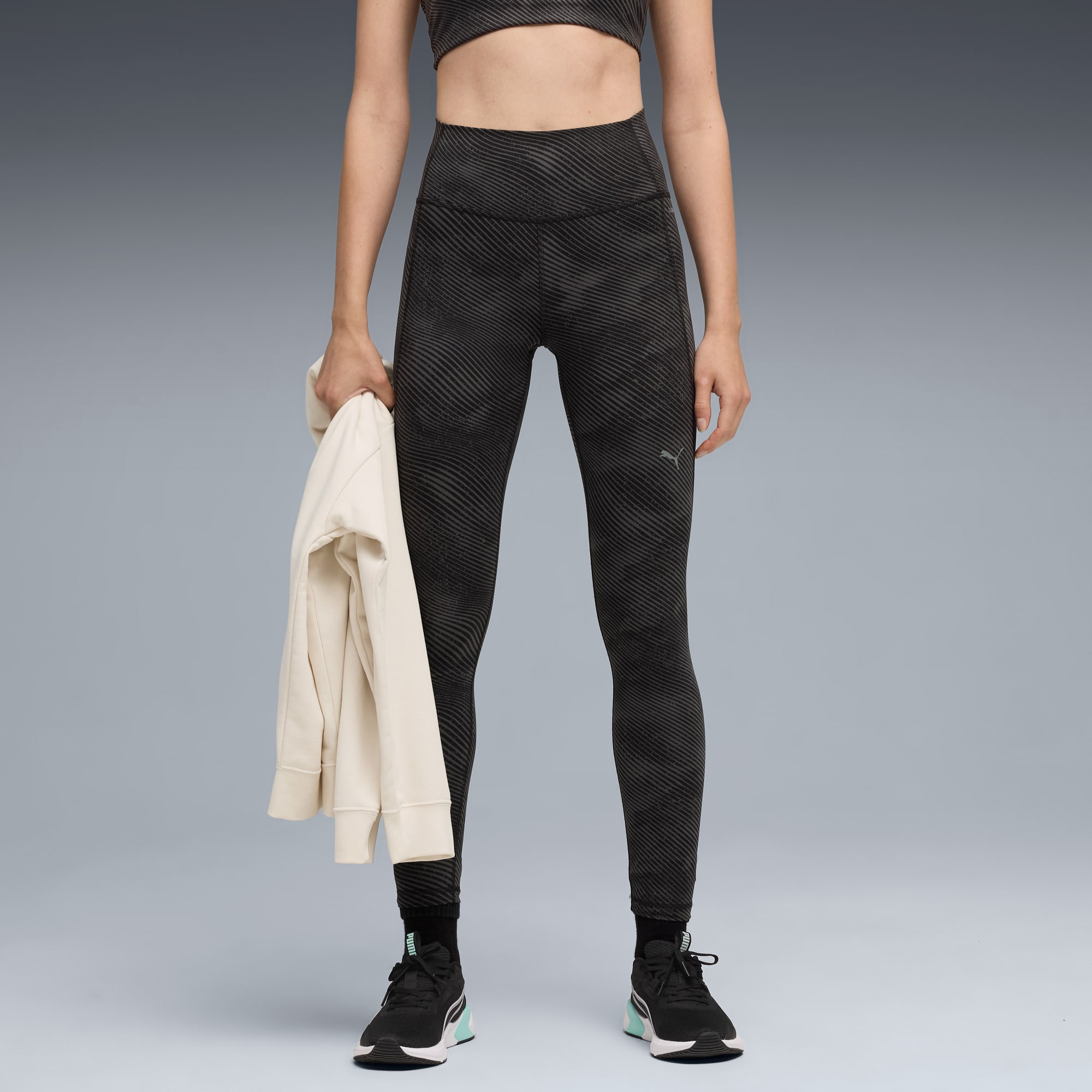 Against grey, a model poses wearing patterned leggings and trainers, holding a jacket.