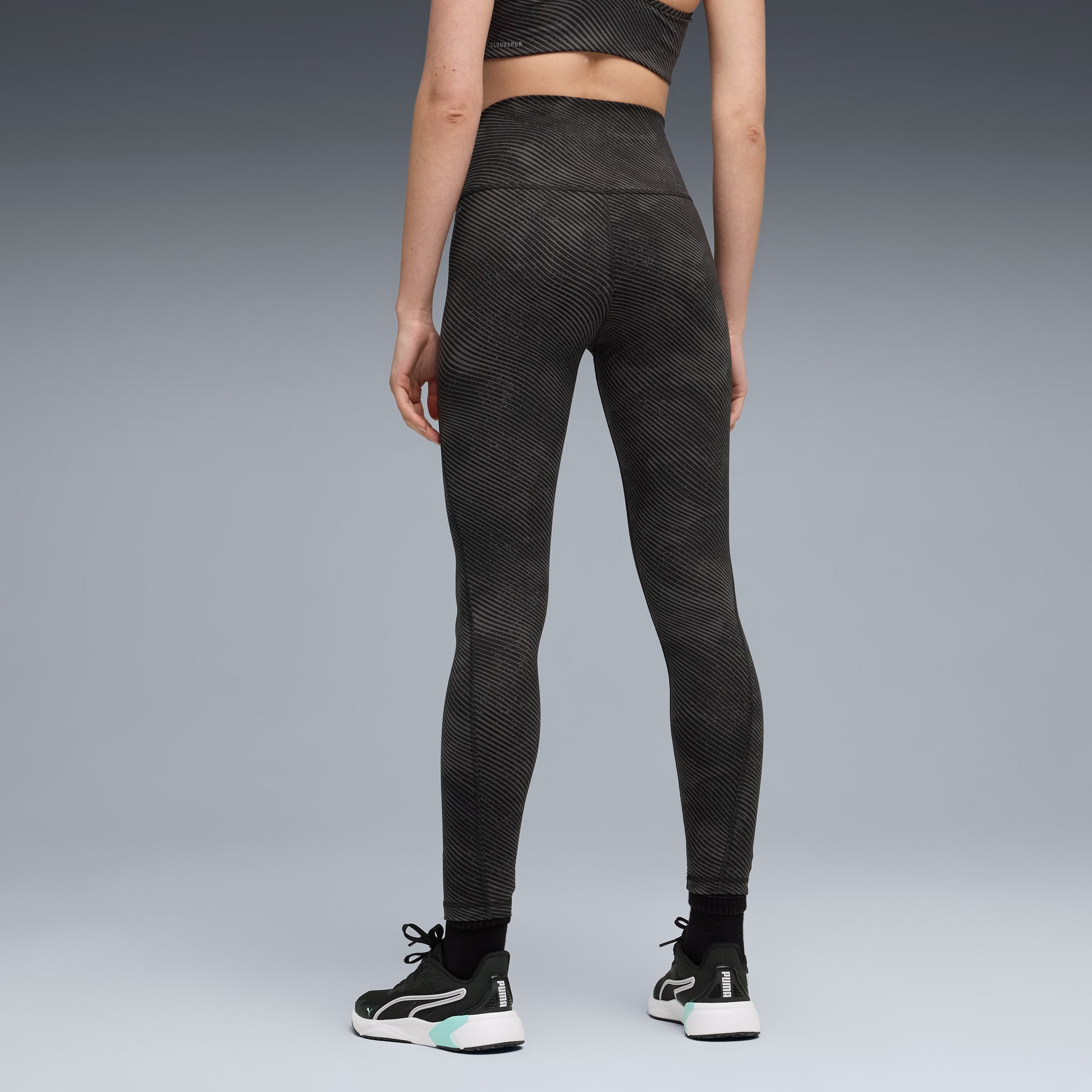 A model faces away on grey, wearing patterned leggings, top and black trainers.