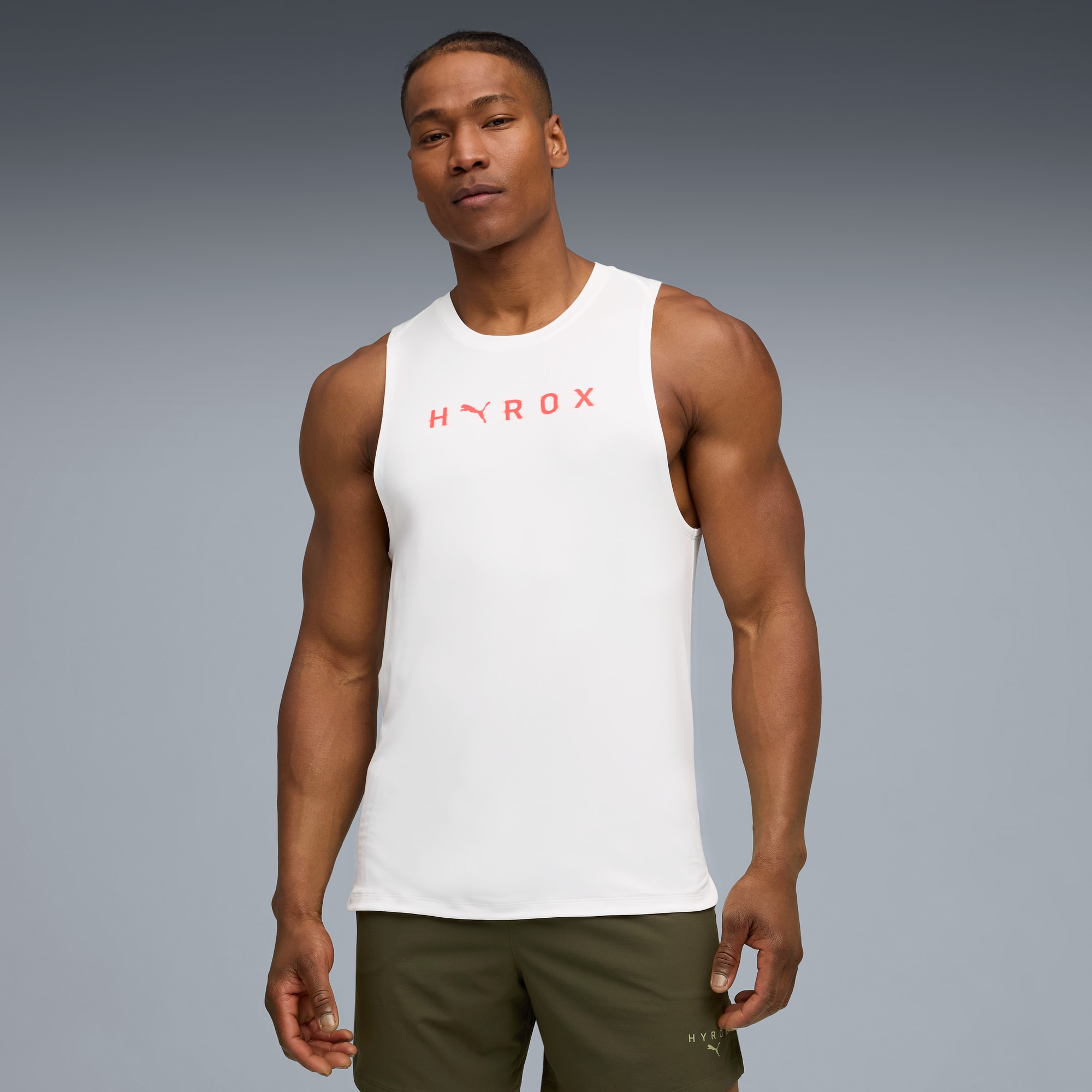 Vor grauem Hintergrund posiert ein Model in einem weißen Top mit rotem Text und olivgrünen Shorts.