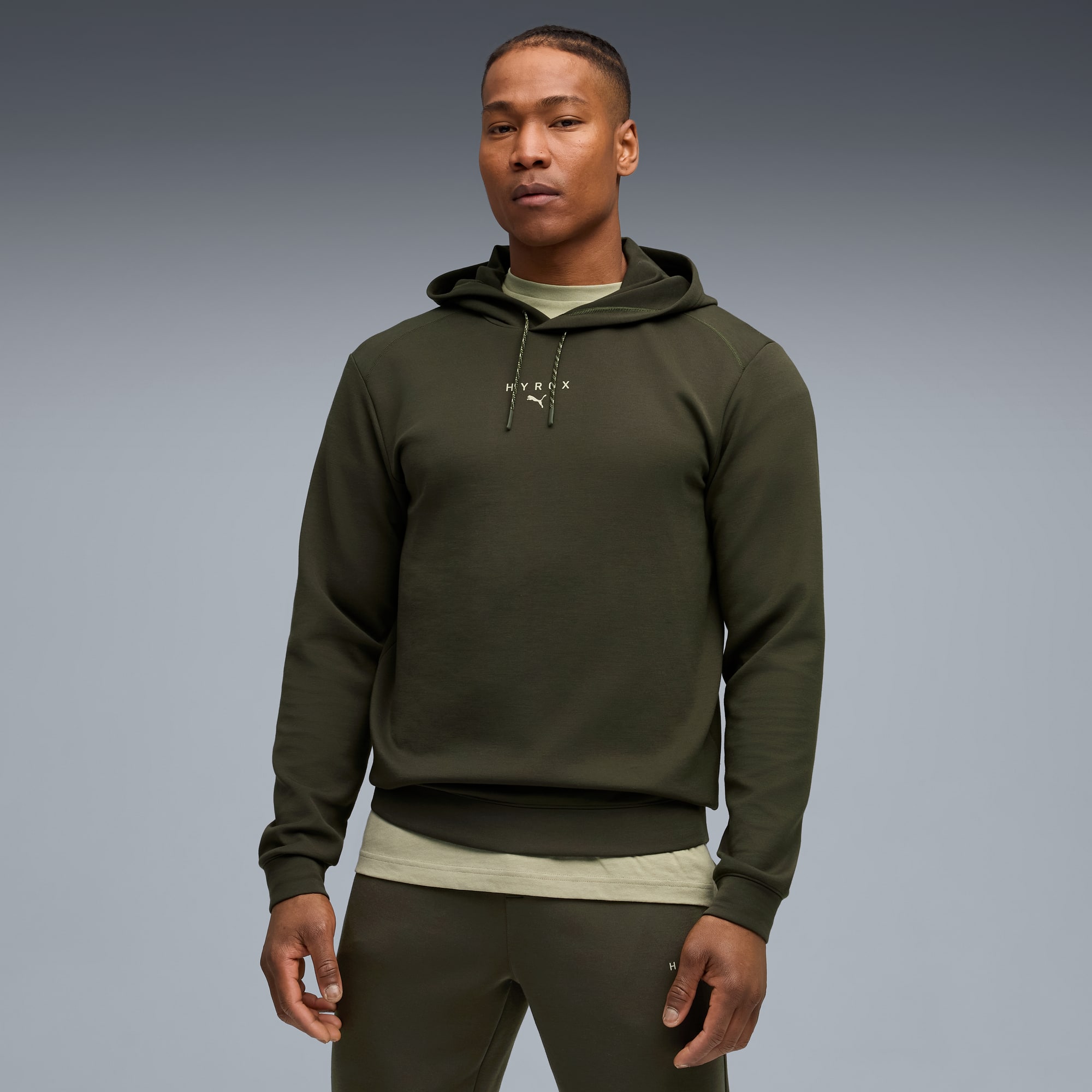 Un modèle pose sur fond gris, portant un sweat à capuche vert foncé et un pantalon de survêtement assorti.