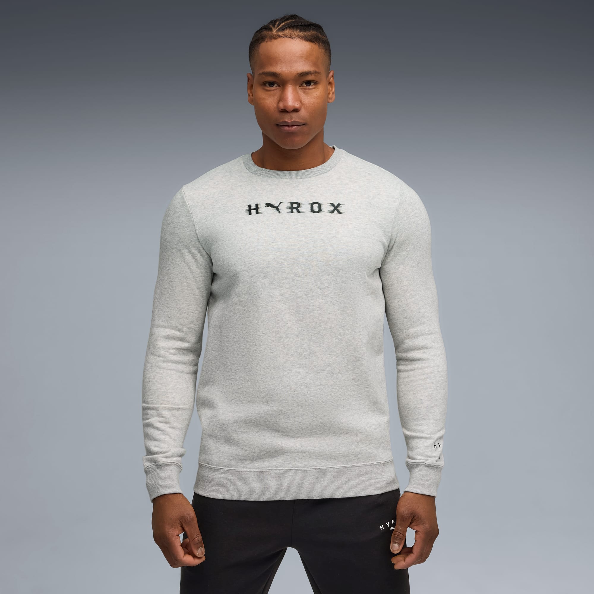 Een model staat tegen een grijze achtergrond, gekleed in een grijze 'HYROX' sweater en zwarte broek.