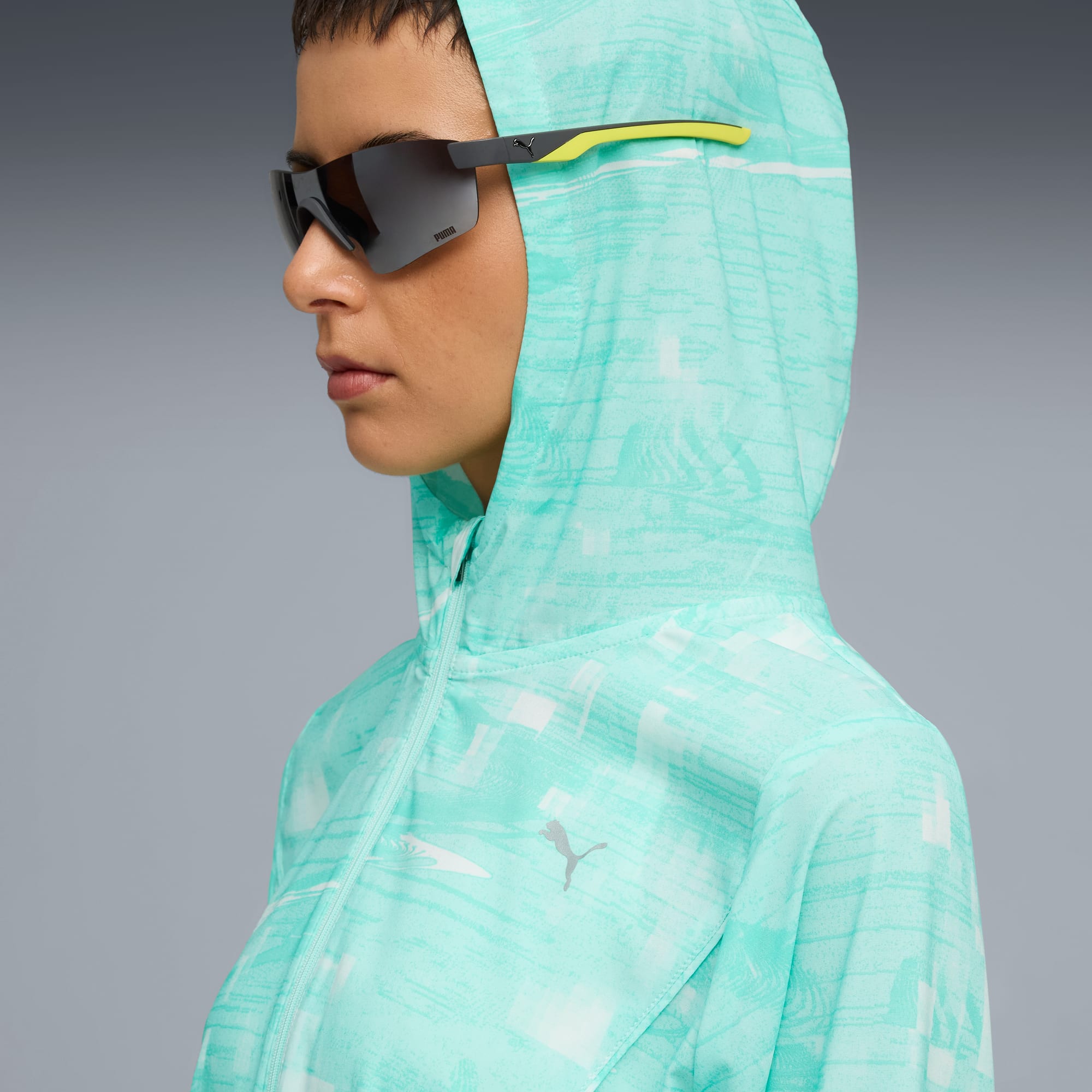 Model trägt Sonnenbrille und gemusterte Kapuzenjacke vor grauem Hintergrund.