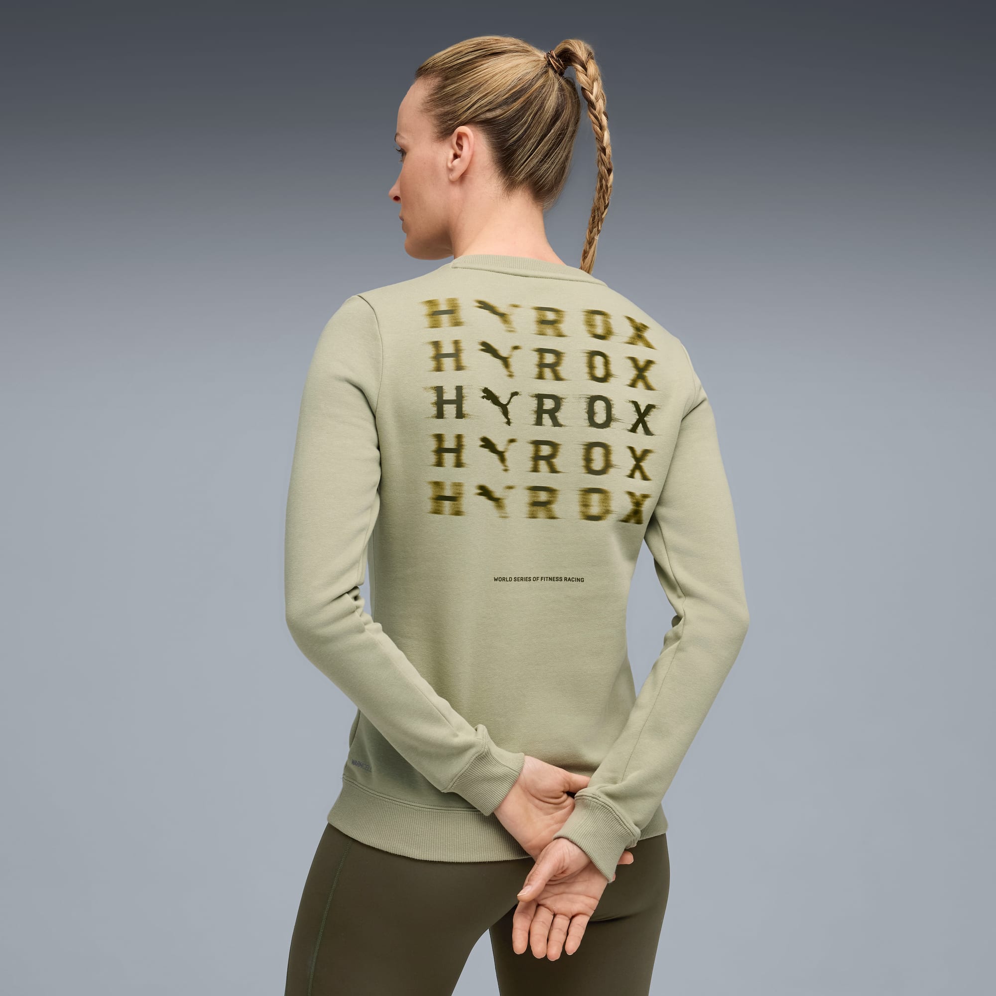 Een model staat met de rug naar de camera op grijs, met een lichtgroen sweatshirt met 'HYROX' erop.