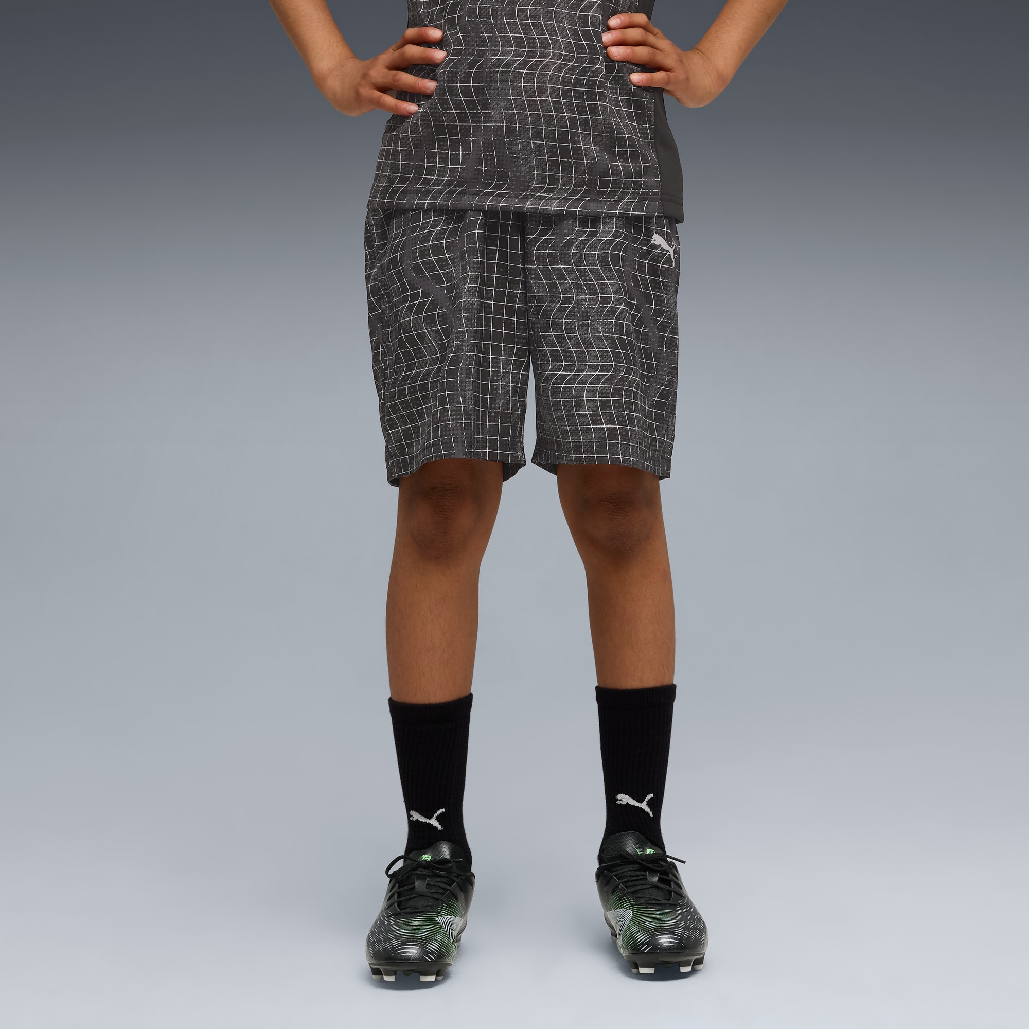 Sur fond gris, un modèle pose en short à motifs, haut assorti, chaussettes noires et chaussures de foot.