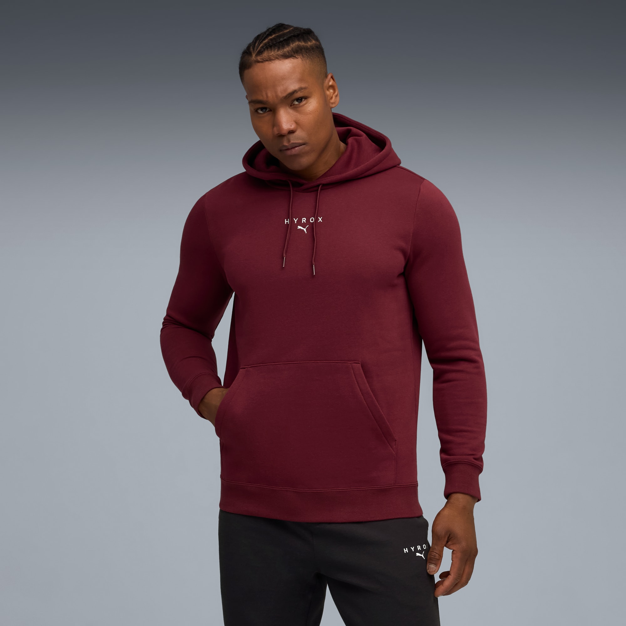 Un mannequin pose sur fond gris, vêtu d'un sweat à capuche bordeaux et d'un pantalon noir.