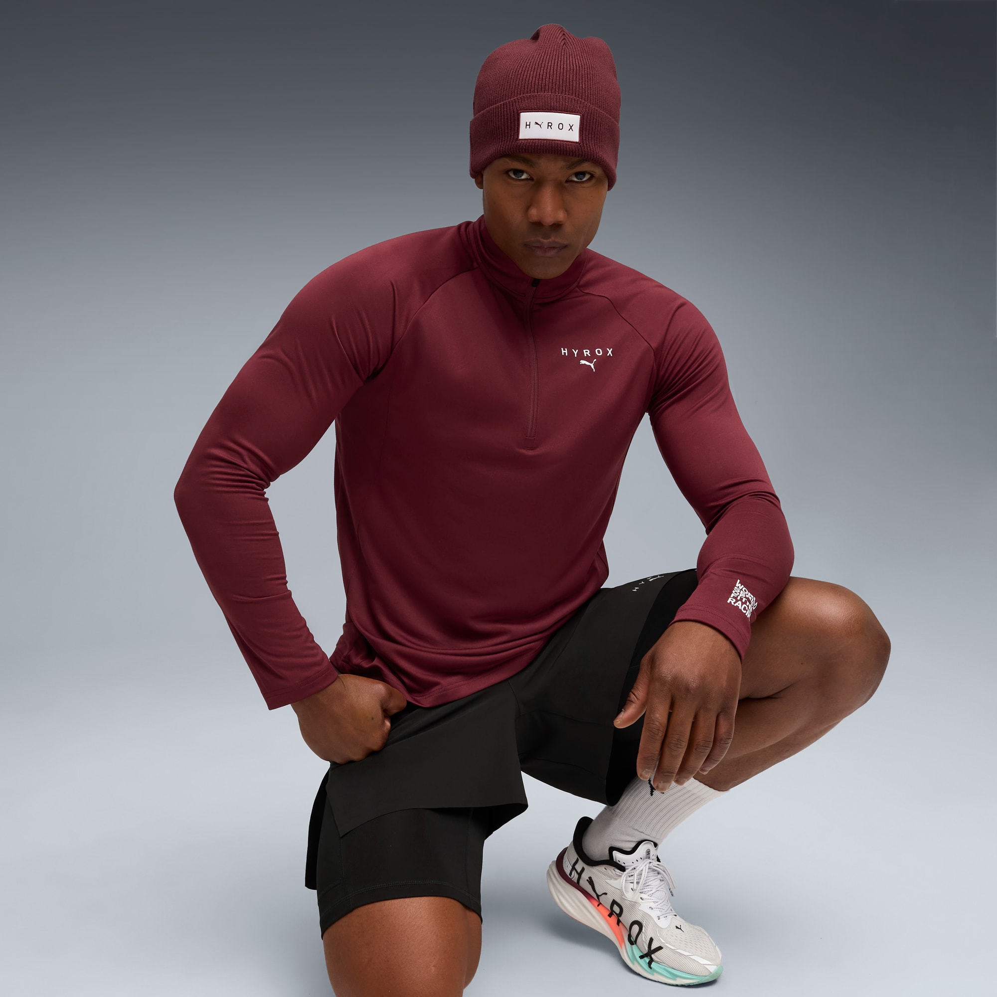 Ein Model vor grauem Hintergrund kauert und trägt Sportbekleidung mit Mütze.
