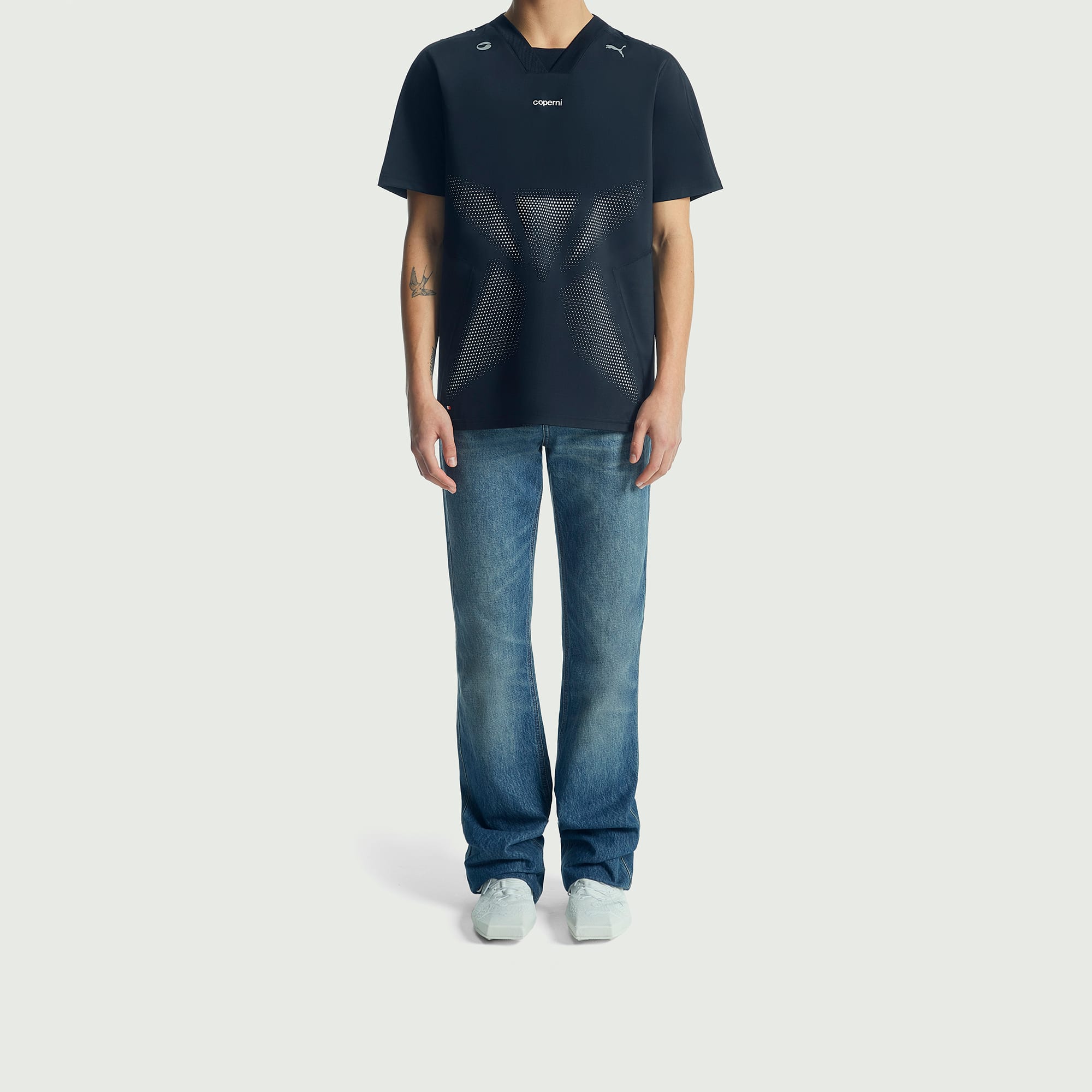 Sur un fond blanc, un mod&egrave;le pose en t-shirt fonc&eacute;, jean et baskets claires.