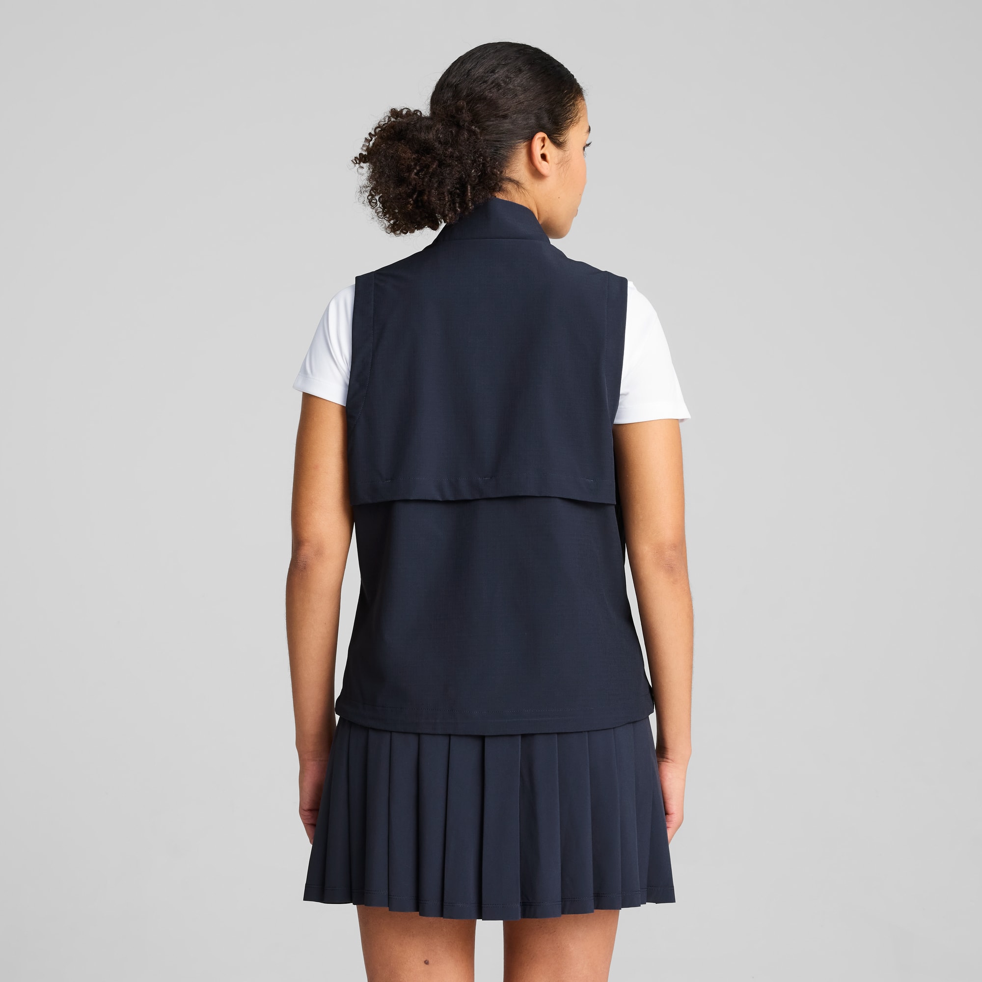 Ein Model vor grauem Hintergrund, mit weißem T-Shirt, marineblauer Weste und Faltenrock.