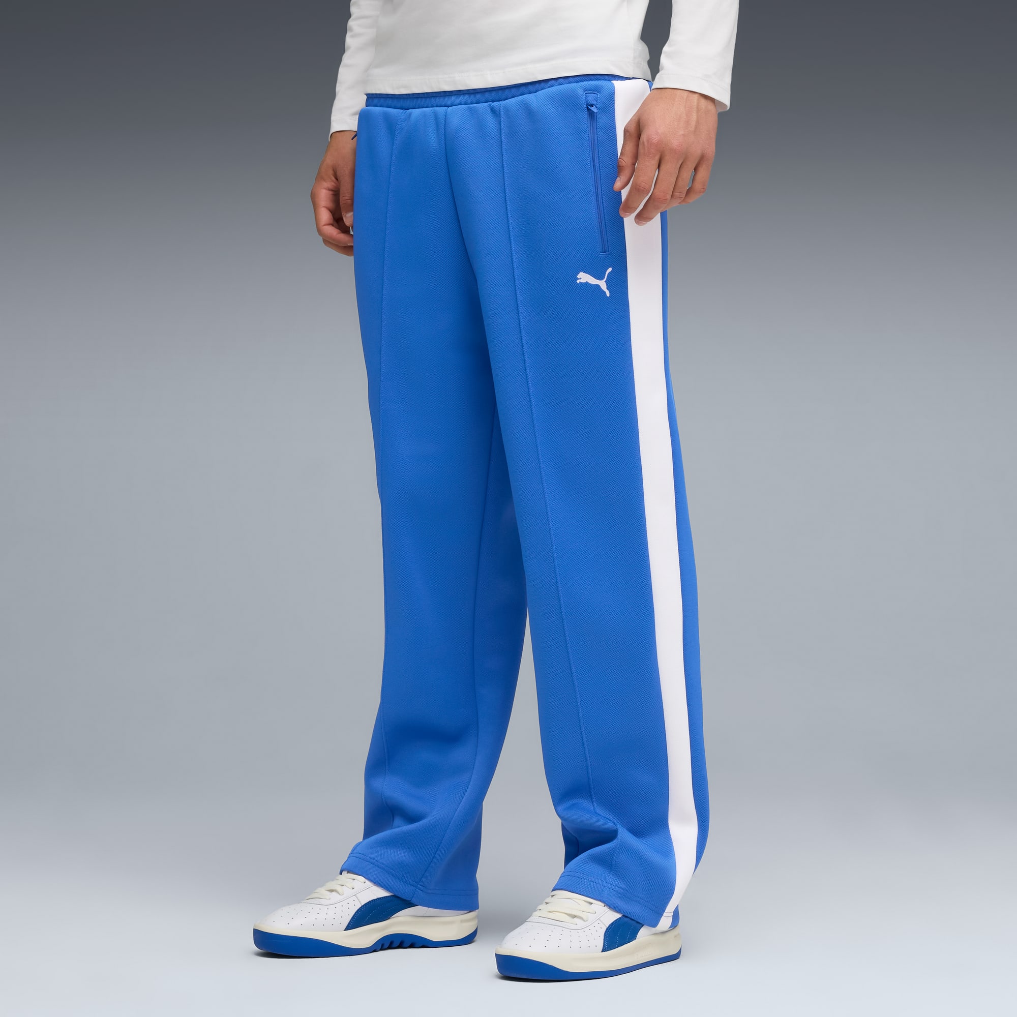 Op een grijze achtergrond draagt een model een blauwe joggingbroek met een witte streep en sportschoenen.