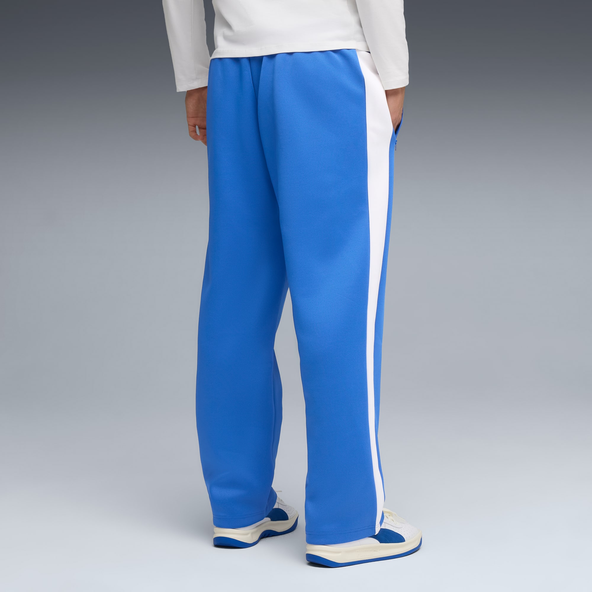 Vor grauem Hintergrund trägt ein Model blaue Hosen mit weißem Streifen und weiße Turnschuhe.