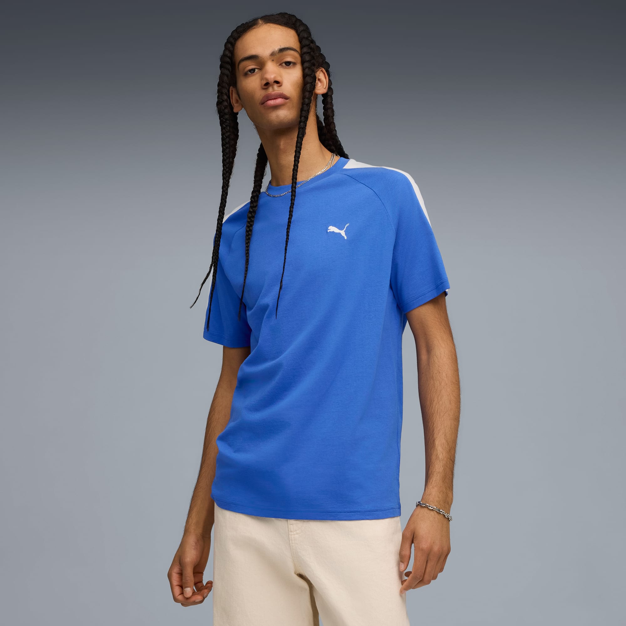 Un modello con trecce posa su sfondo grigio con t-shirt blu e pantaloncini color crema.