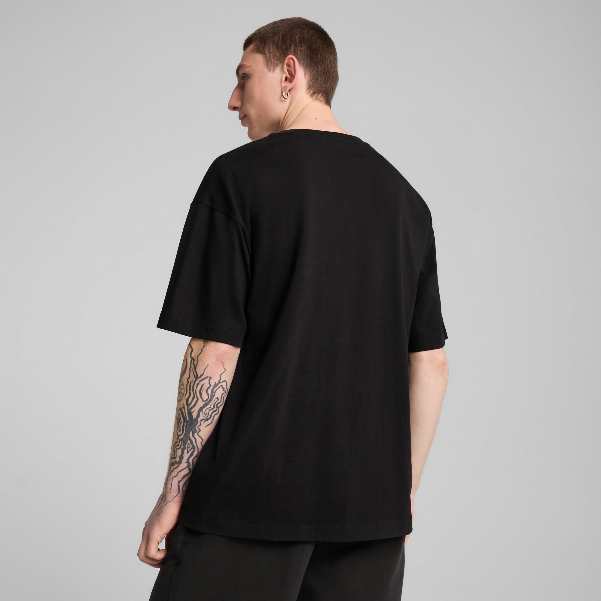 Eine Person mit Tattoo am Arm, vor grauem Hintergrund, trägt ein schwarzes T-Shirt.