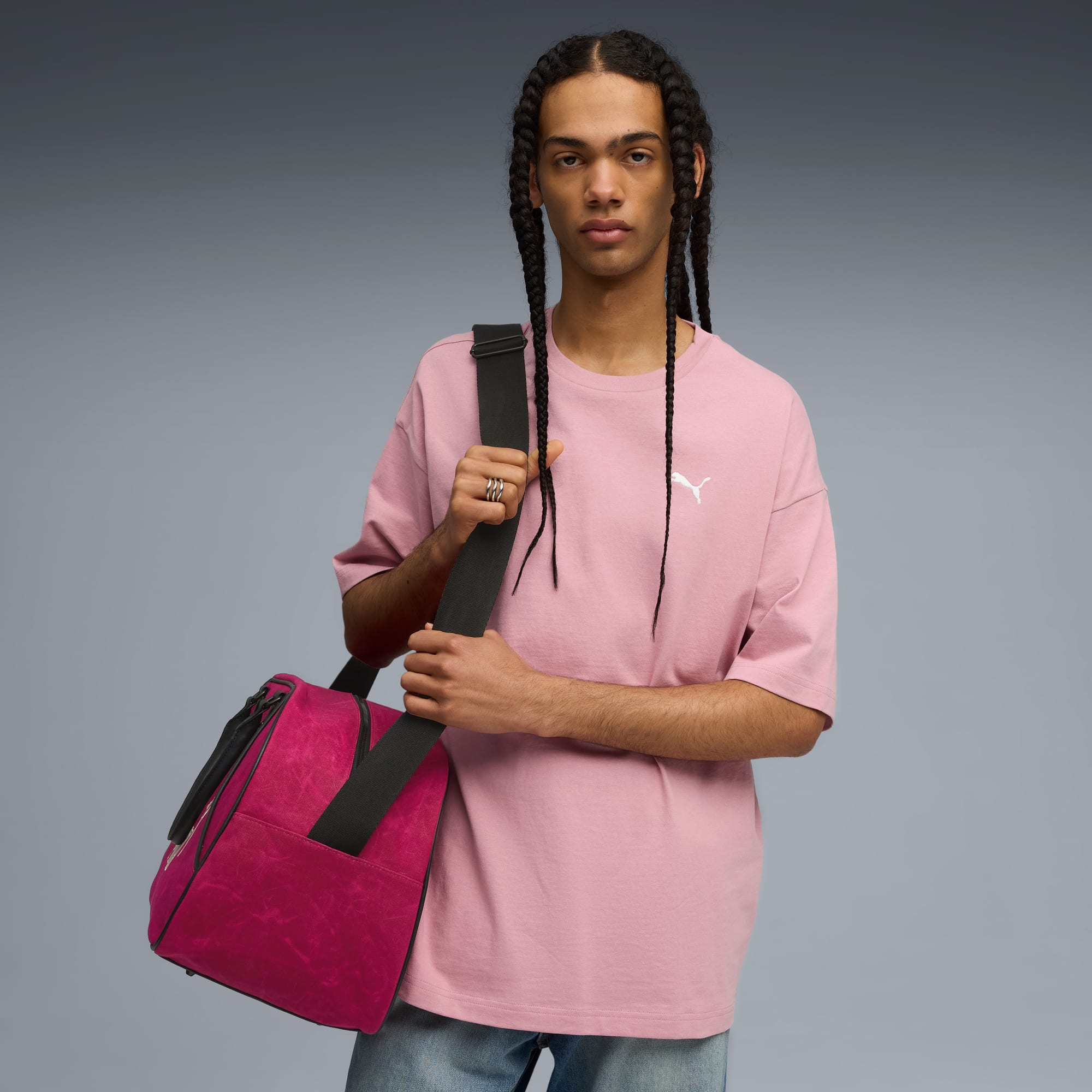 Su sfondo grigio, modello con trecce indossa t-shirt rosa e porta borsone rosa.