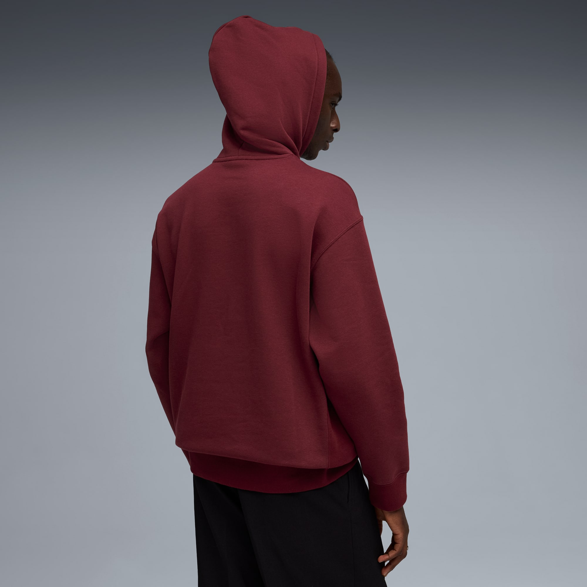 Een model poseert tegen een grijze achtergrond, gekleed in een bordeauxrode hoodie en zwarte broek.