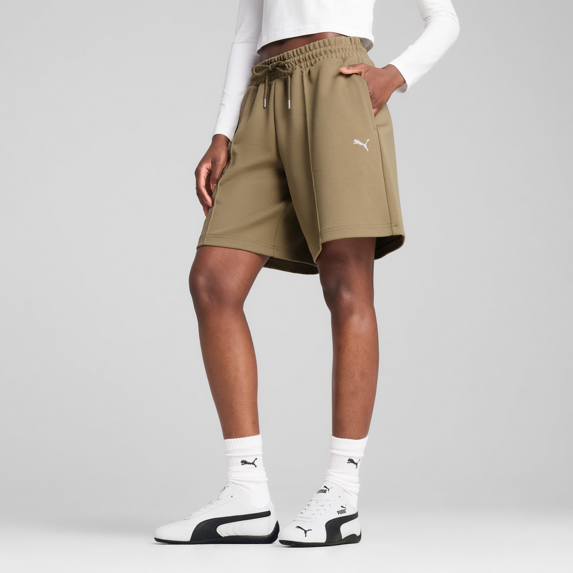 Tegen een grijze achtergrond poseert een model in witte top, kaki shorts en sportschoenen.