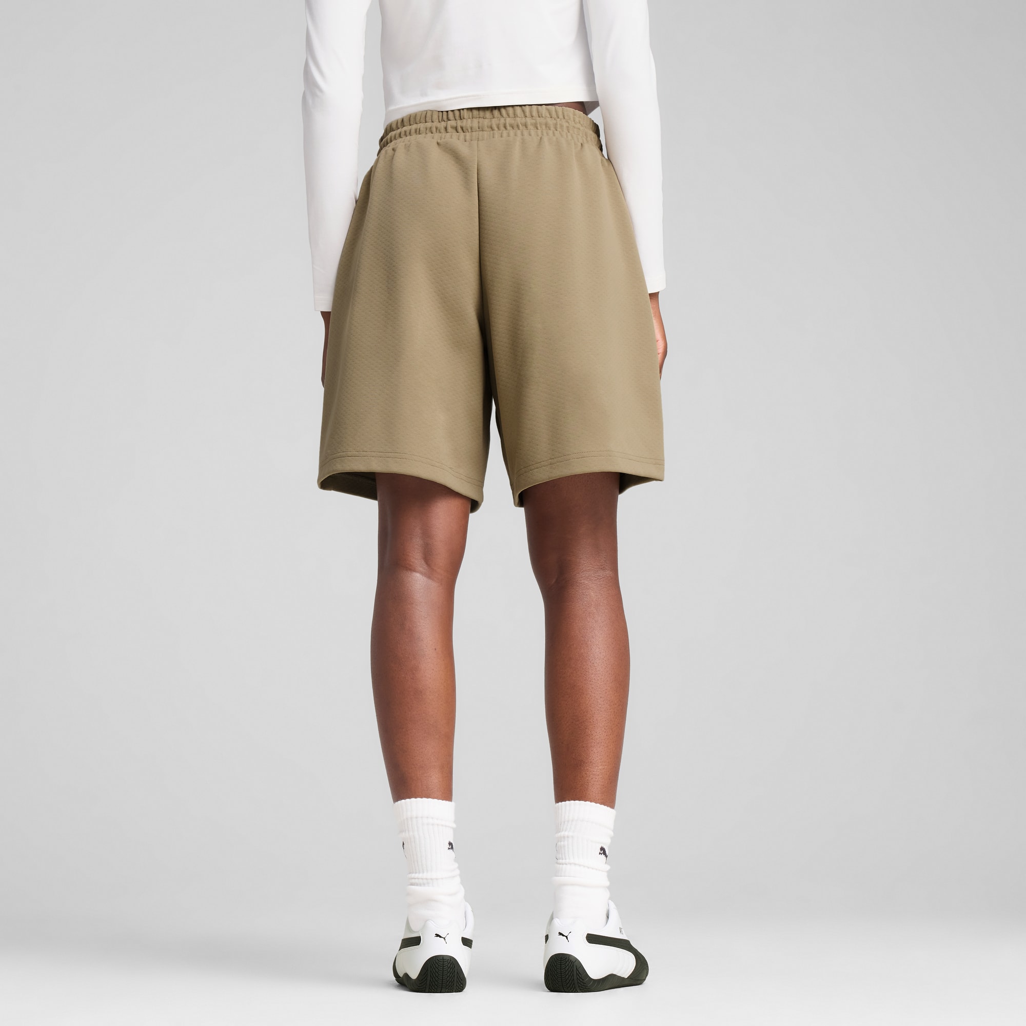 Een model poseert tegen een grijze achtergrond, met kaki shorts, witte top, sokken en sneakers.