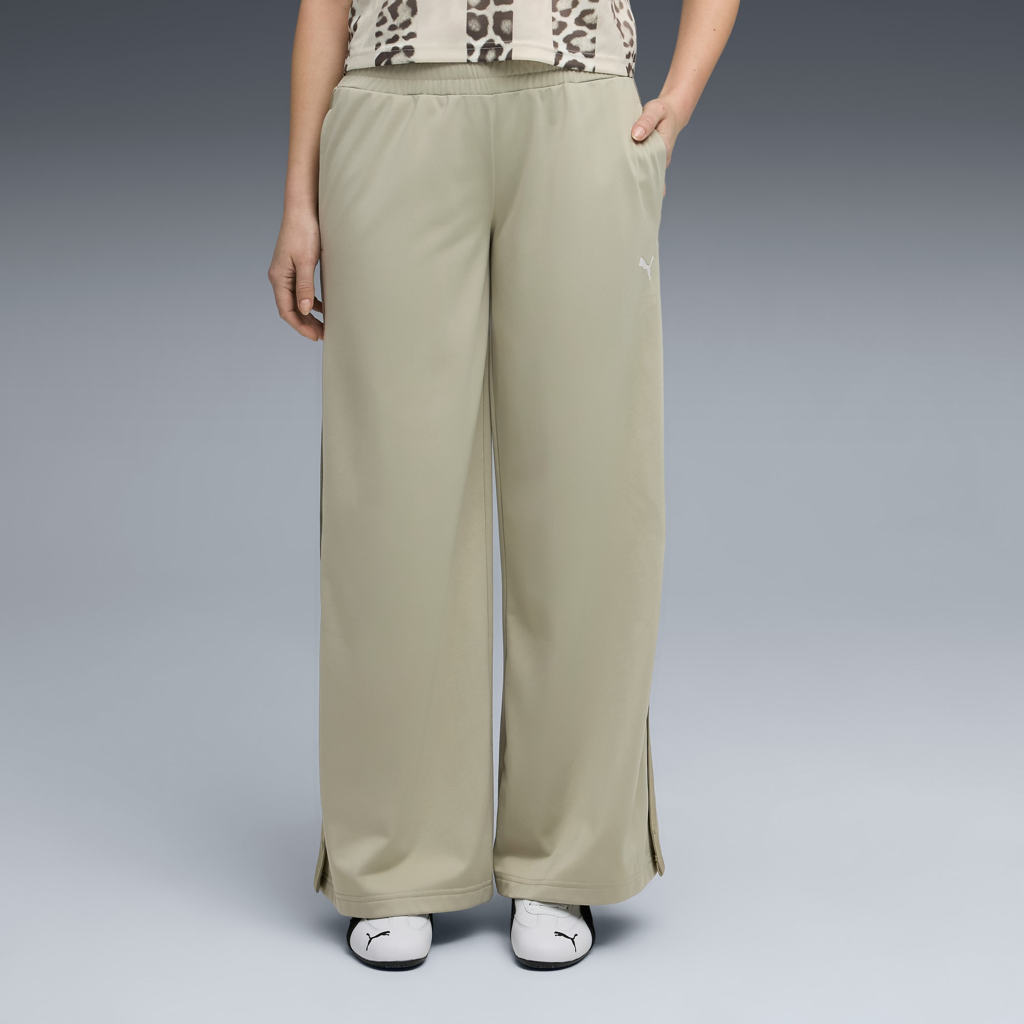 Un mannequin pose sur fond gris, portant un pantalon beige, un haut à motifs et des chaussures blanches.