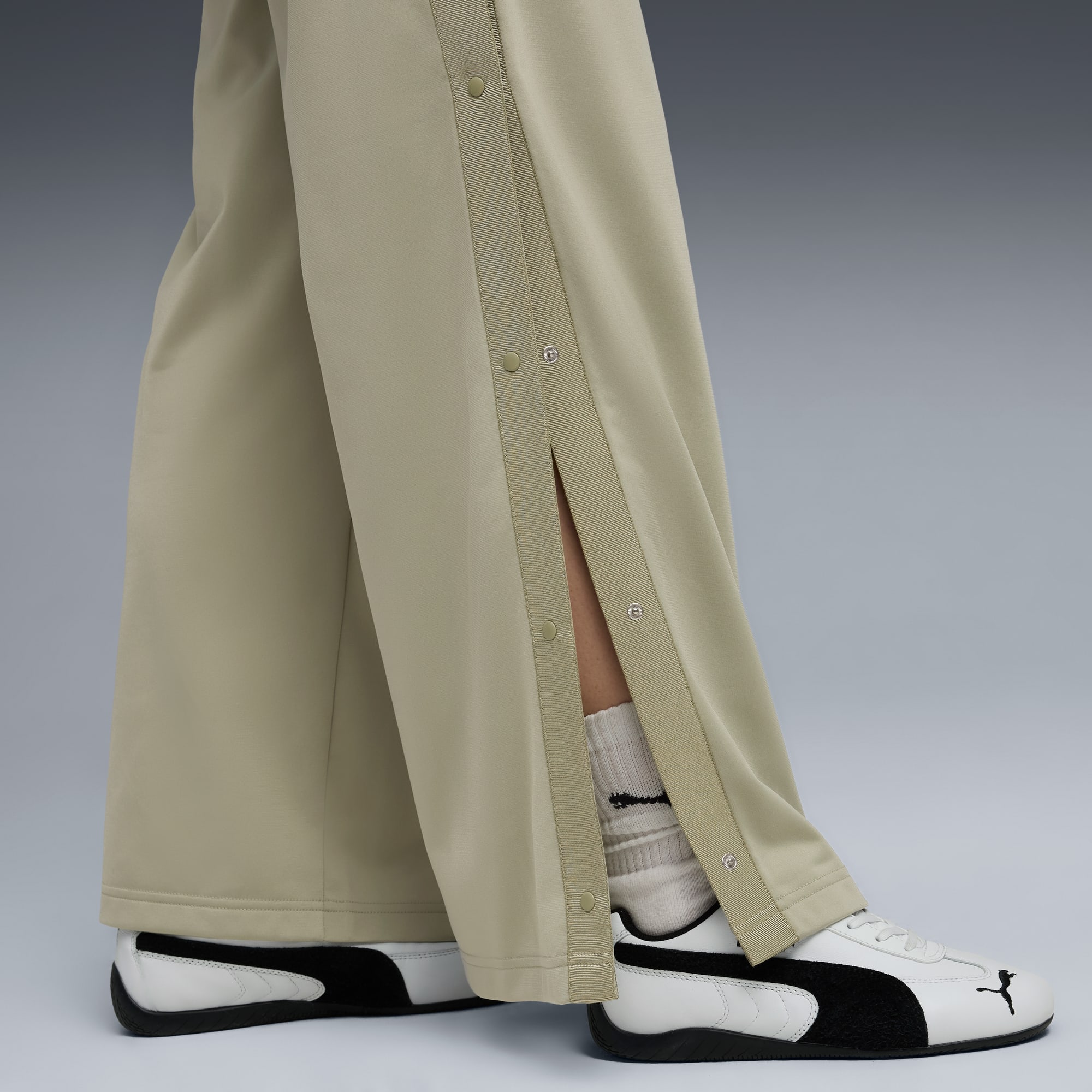 Sur fond gris, jambes en pantalon beige à boutons-pression, chaussettes et chaussures noir et blanc.