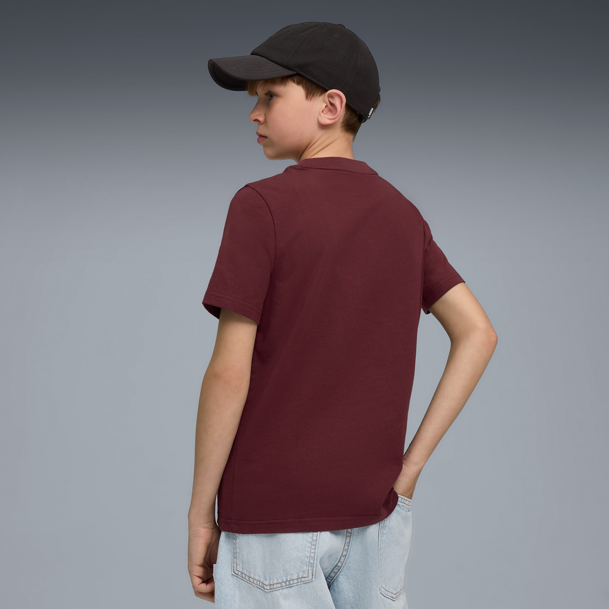 Een kind poseert tegen een grijze achtergrond met een pet, bordeauxrood T-shirt en jeans.