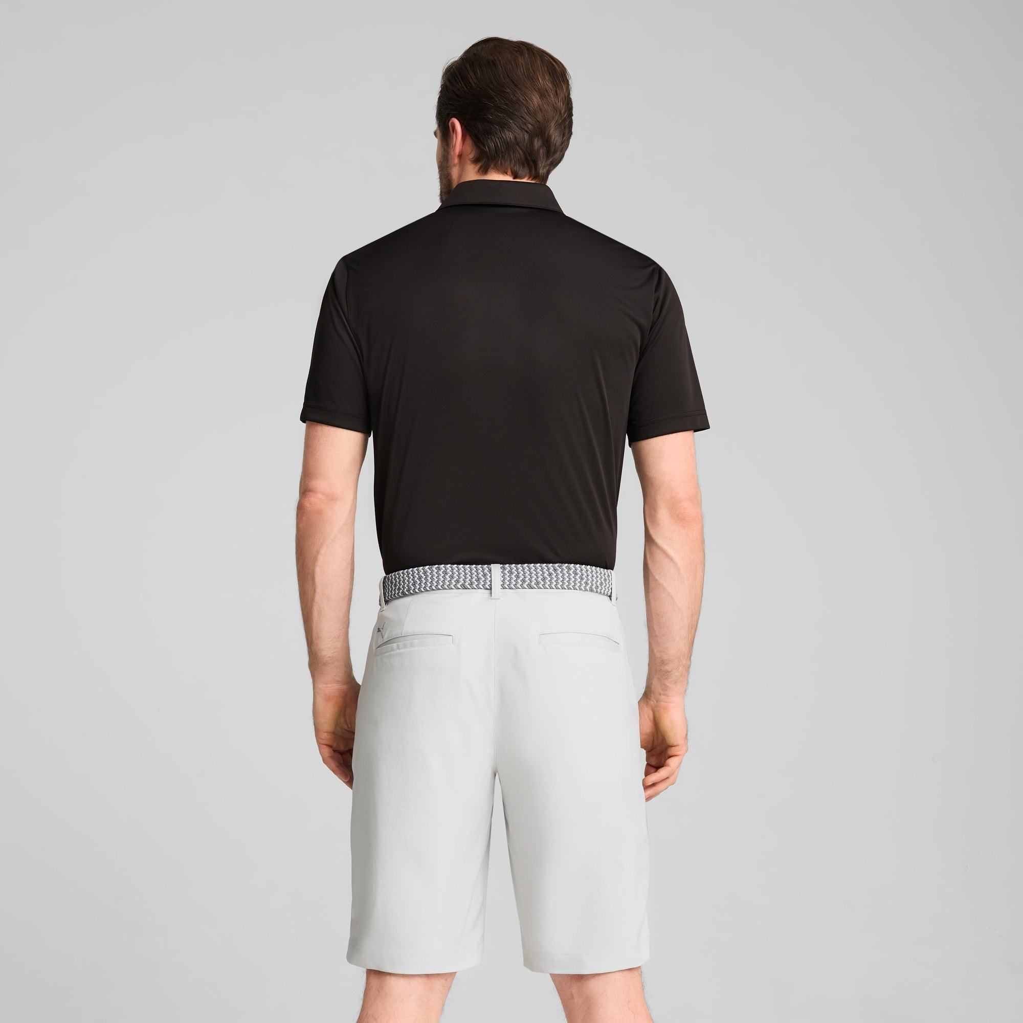 Een model staat met de rug naar de camera op grijs, draagt zwarte polo en lichtgrijze shorts met riem.