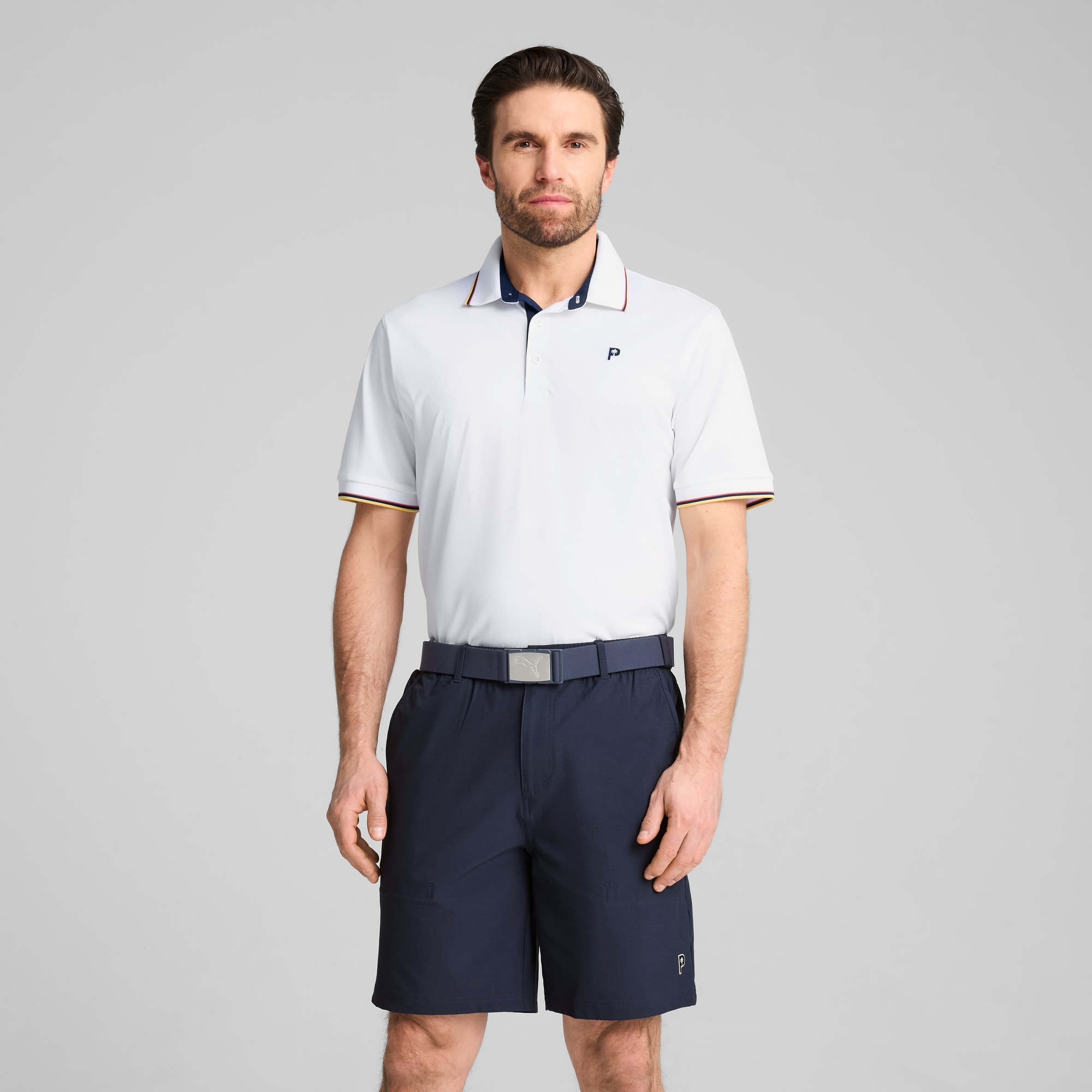Tegen een grijze achtergrond staat een model met een wit poloshirt en een marineblauwe short.