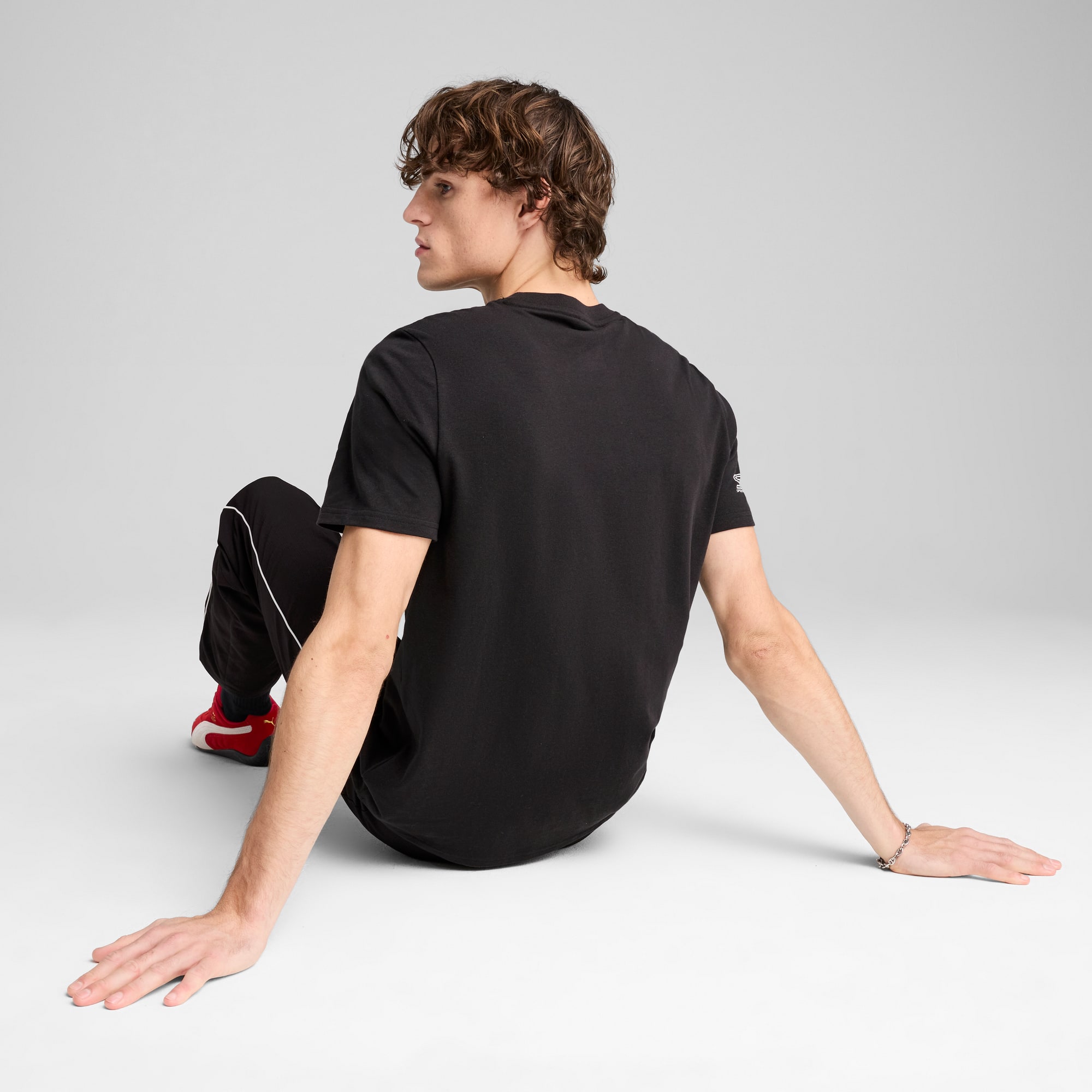 Sur un fond gris, un modèle est assis portant un t-shirt noir, un pantalon de survêtement et des baskets rouges.