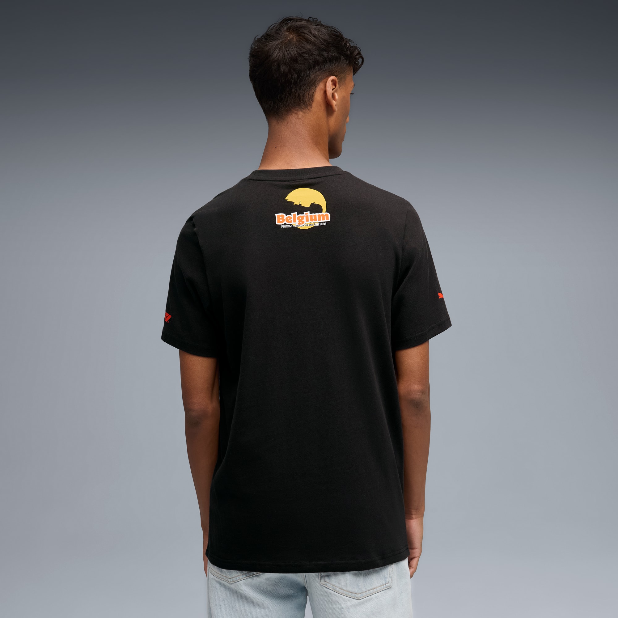 Un modello di spalle su sfondo grigio, indossa una t-shirt nera con grafica e jeans chiari.