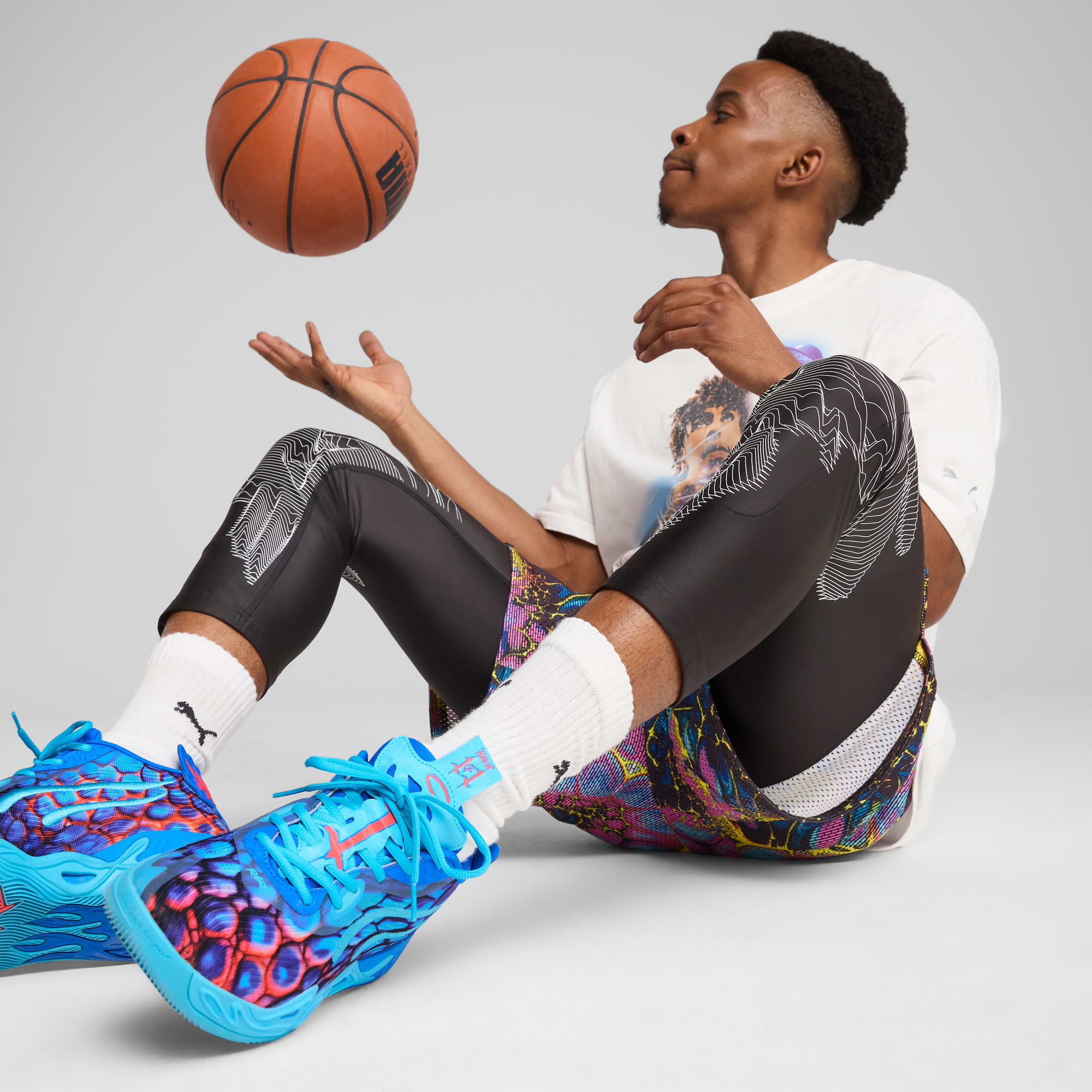 Op grijs zit een model te jongleren met een basketbal, gekleed in een t-shirt, korte broek en blauwe sportschoenen.