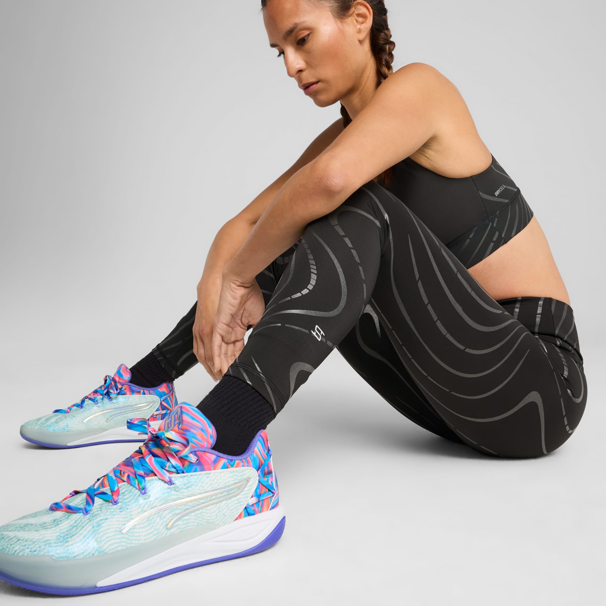 Ein Model sitzt vor einem grauen Hintergrund und trägt gemusterte schwarze Sportkleidung und bunte Turnschuhe.