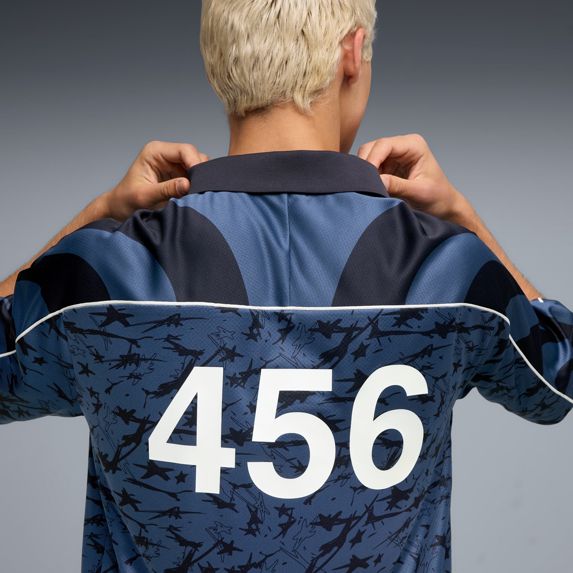 Una persona sistema una maglia blu fantasia con il numero 456 sul retro, su sfondo grigio.